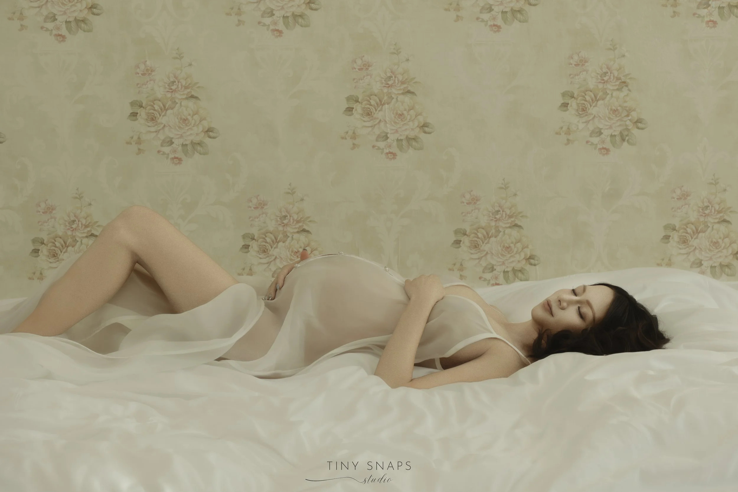 A pregnant woman in a sheer white dress lying on a bed with floral wallpaper in the background, smiling with her eyes closed.