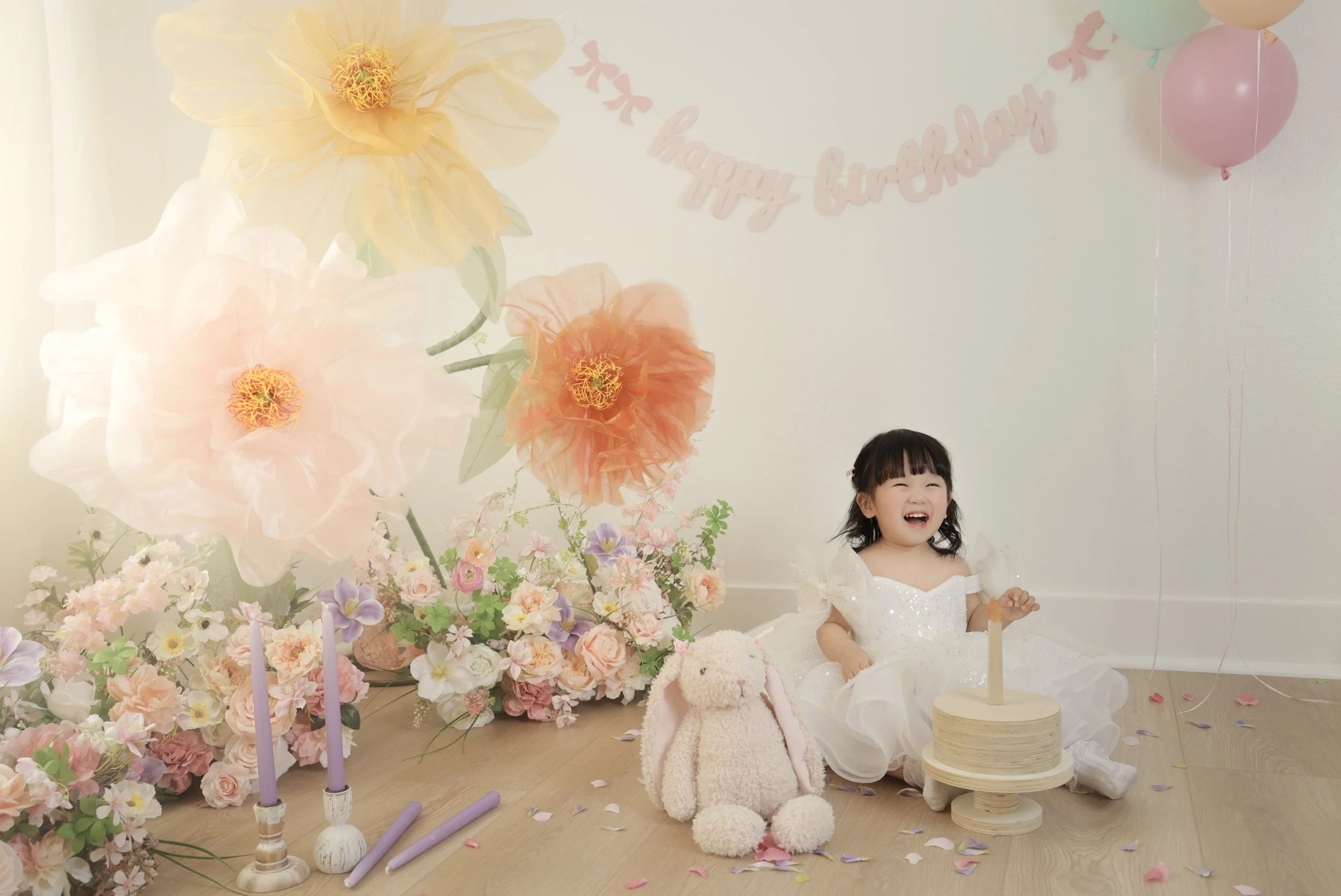 A young girl in a white dress celebrates her birthday with a small cake, surrounded by pastel-colored balloons, flowers, and decorations. She is sitting on the floor, smiling happily, with a plush bunny toy nearby. The background features a birthday 
