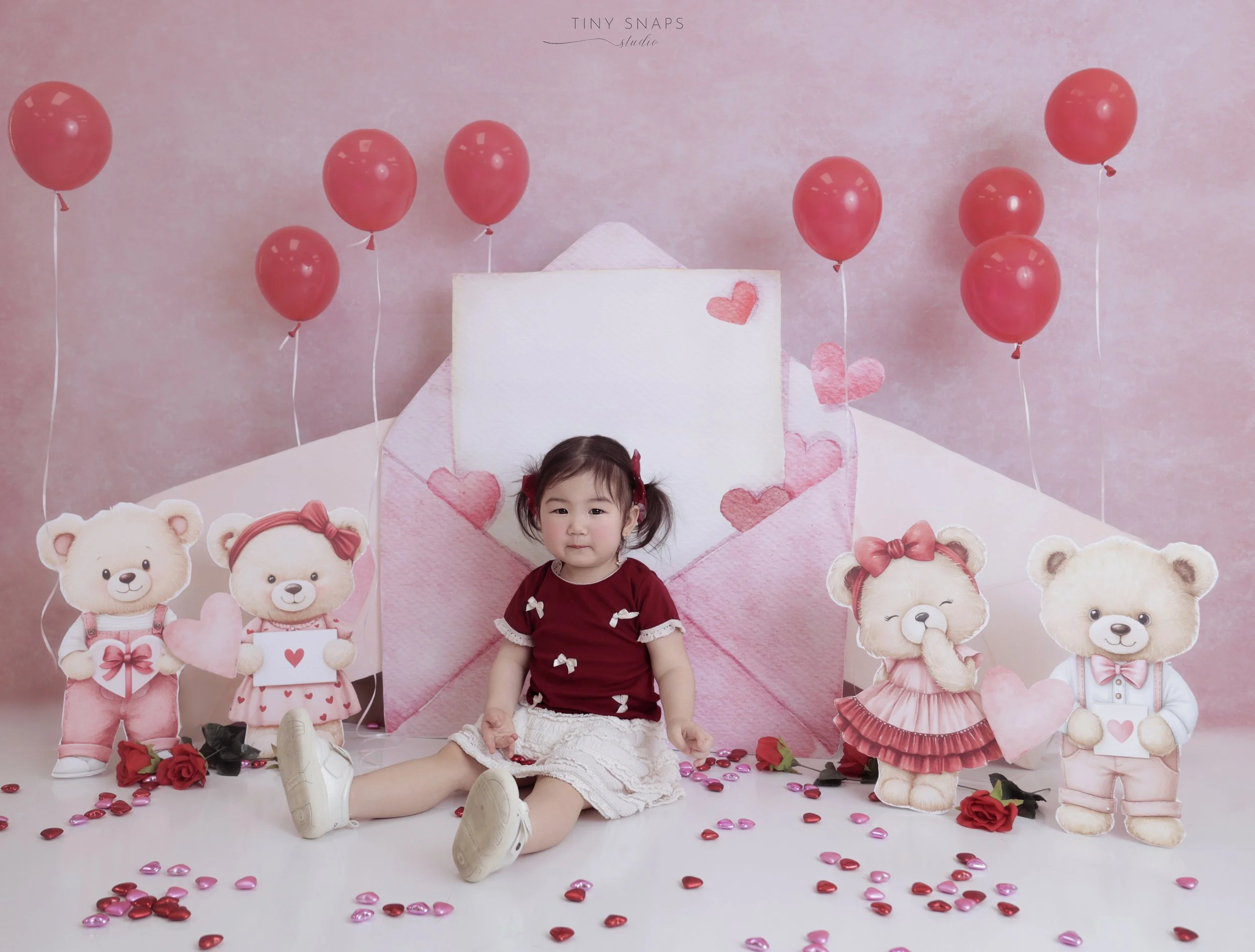 A young girl sitting on the floor in front of a pink Valentine's Day-themed backdrop with paper teddy bears, balloons, and heart-shaped decorations.