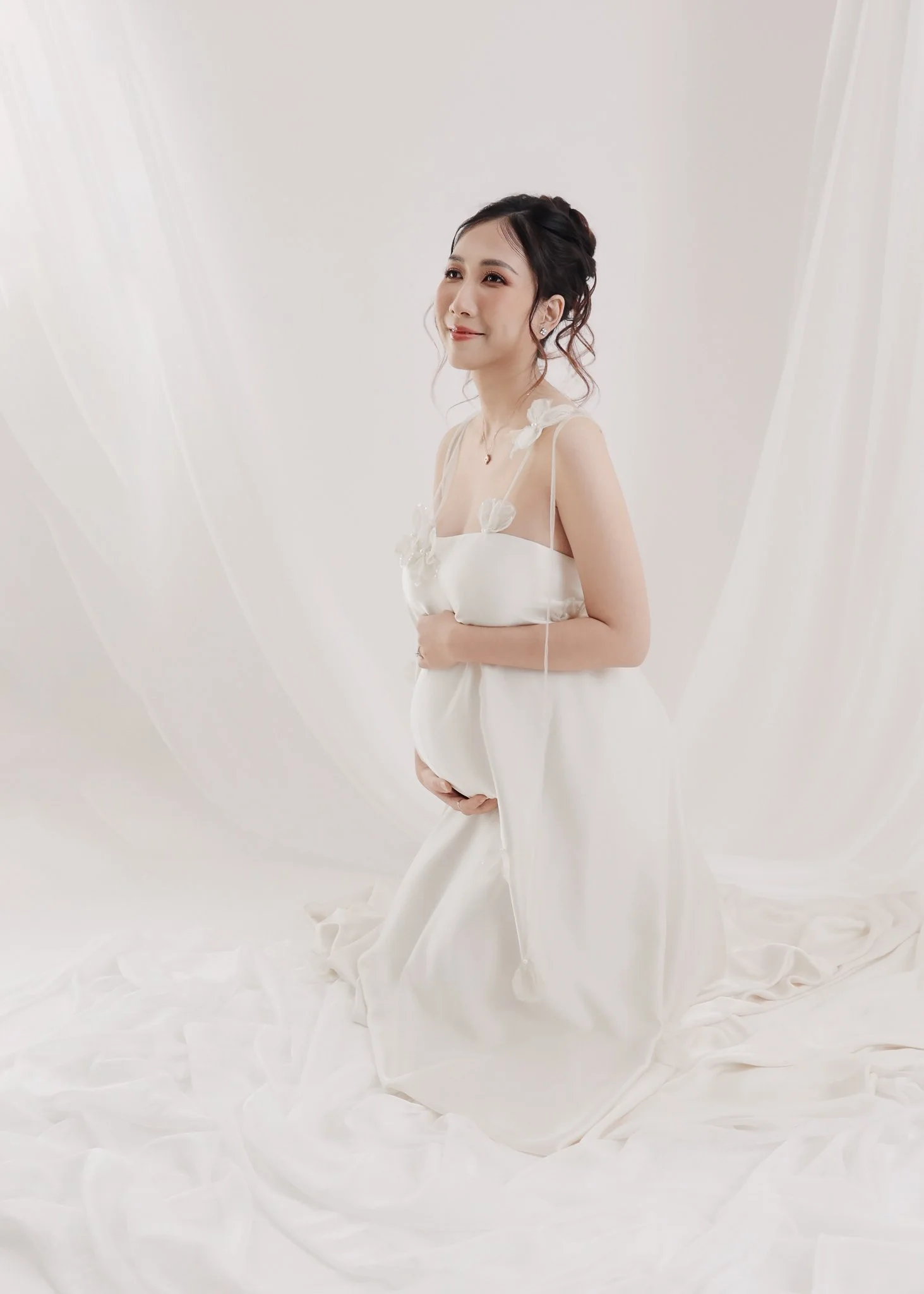 A pregnant woman in a white dress, standing on a white fabric backdrop, with her hand on her baby bump, smiling softly.
