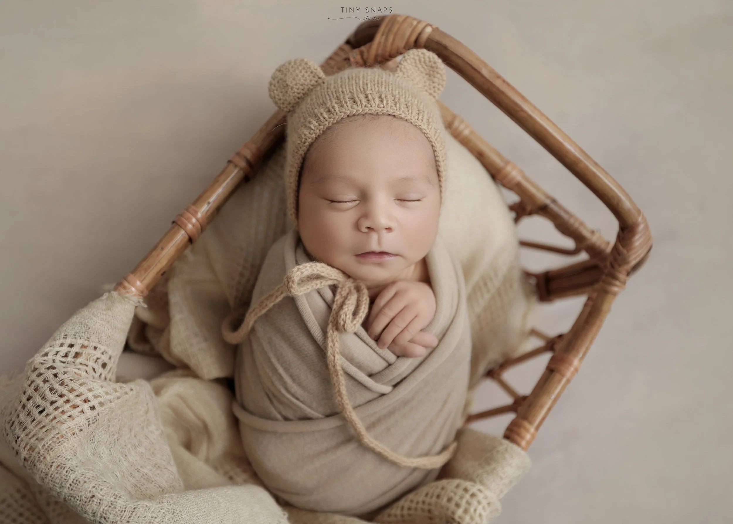 A sleeping baby in a beige knit hat with bear ears, wrapped in a beige blanket, lying in a wicker basket lined with soft fabric and surrounded by cozy blankets.