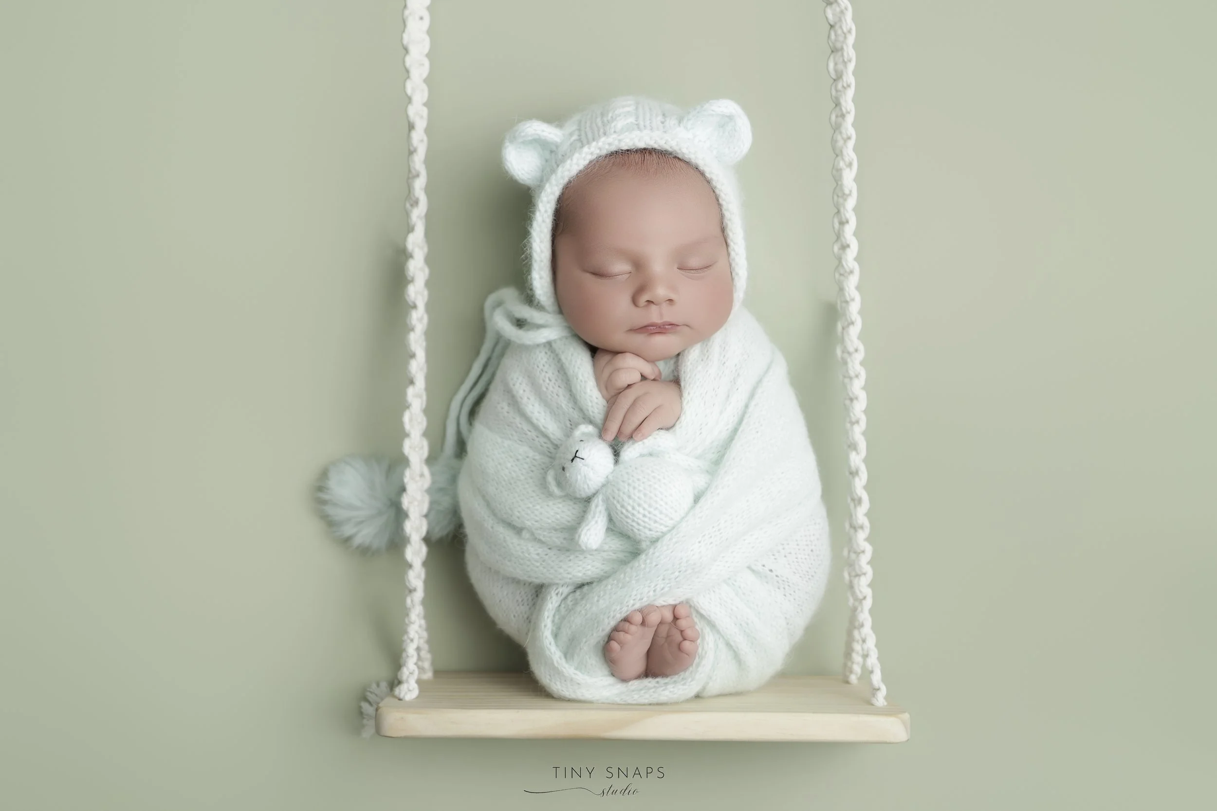 A sleeping baby dressed in white, wearing a knit bear hat, swaddled in a cozy white blanket, on a wooden swing with braided ropes, against a light green background.