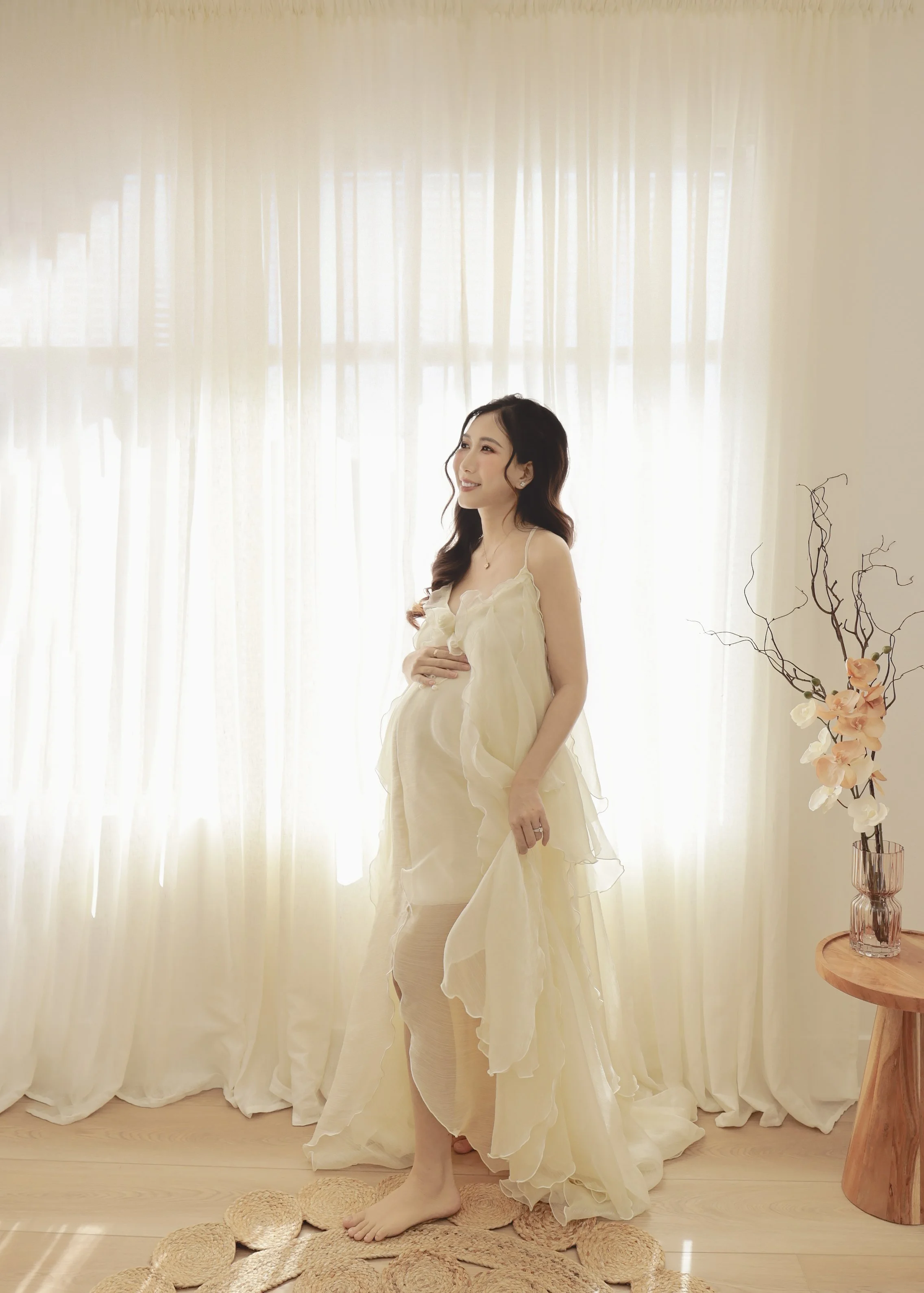 A pregnant woman in a flowing, cream-colored dress stands in a bright room with sheer curtains and a wooden side table with flowers in a glass vase.