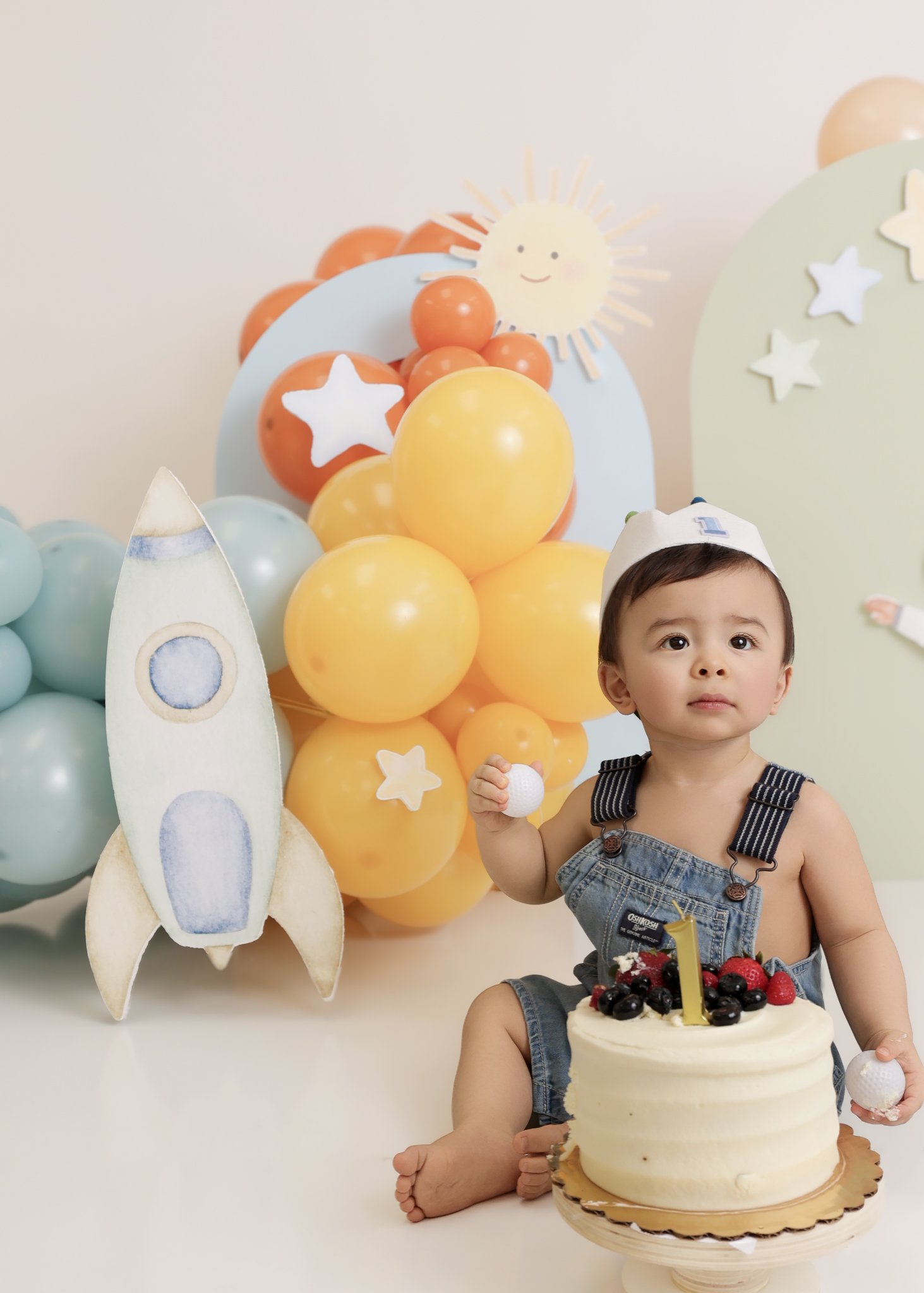 A young boy sitting on the floor holding a piece of cake and a golf ball, celebrating his first birthday with a cake, balloons, and space-themed decorations in the background.