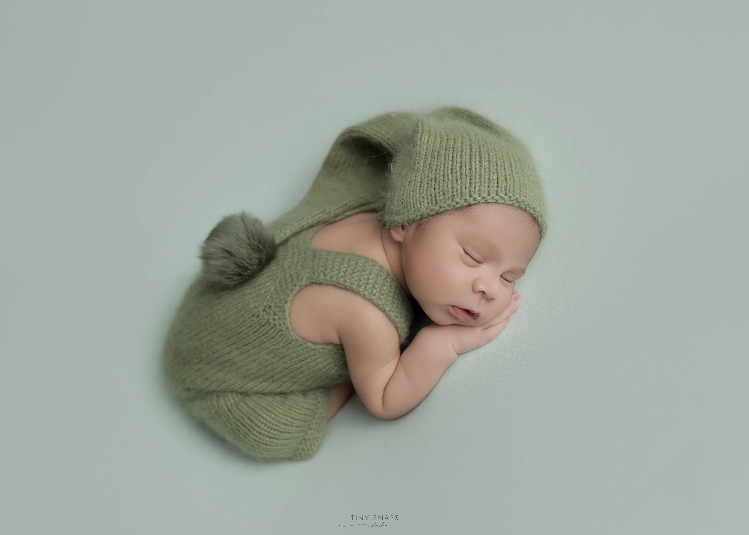 A baby in a green knitted outfit with a matching hat and pom-pom, sleeping on a light green background.
