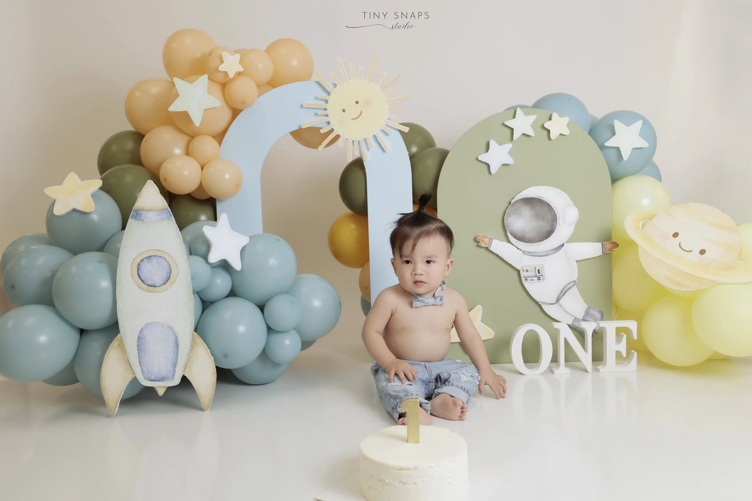 Young boy sitting on the floor in front of space-themed decorations including balloons, a paper rocket, stars, a smiling sun, and an astronaut figure. There is a birthday cake with a number one candle in front of him.