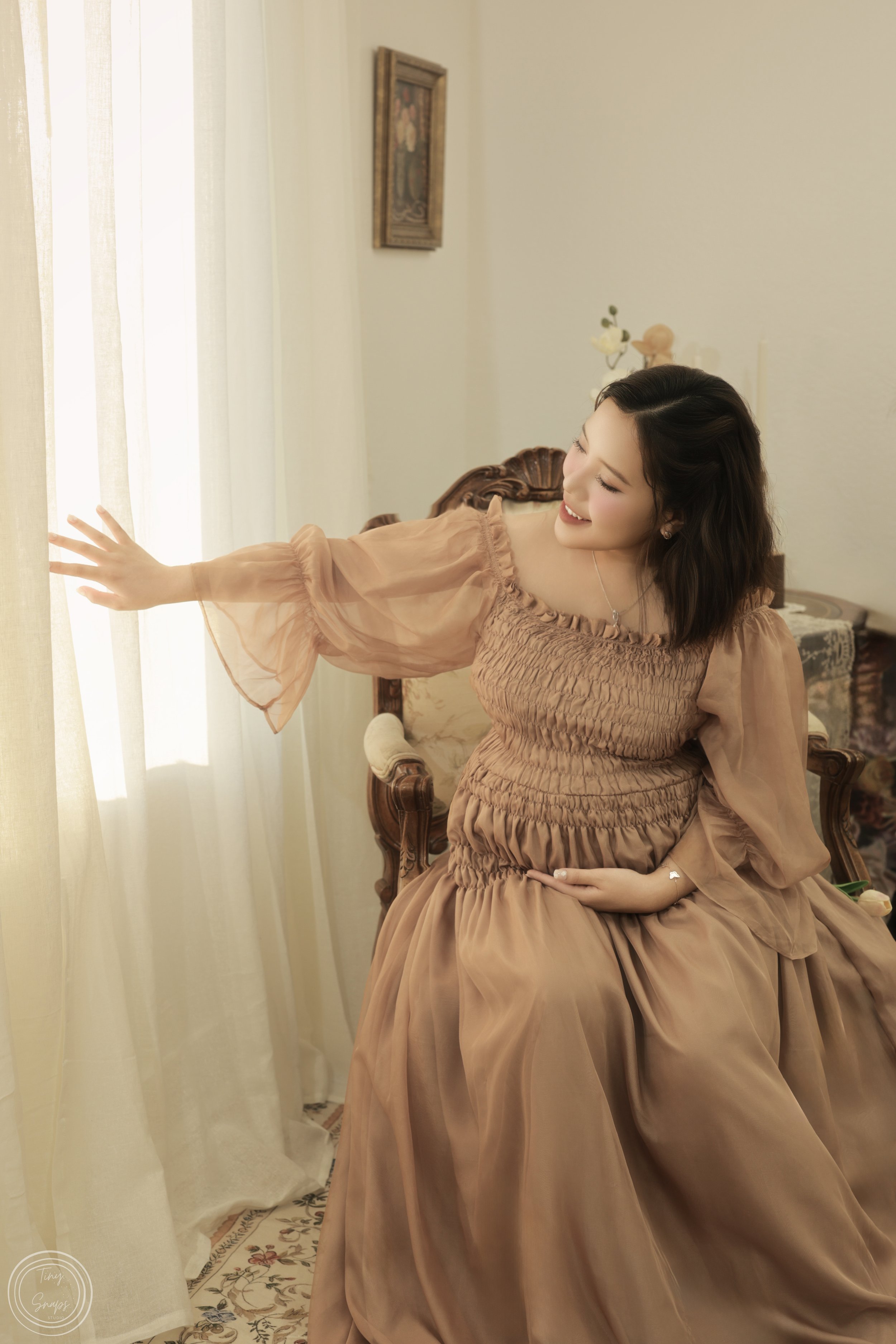 A woman sitting on a vintage wooden chair, smiling and reaching out to touch sheer white curtains, wearing a flowing peach-colored dress, in a softly lit room.