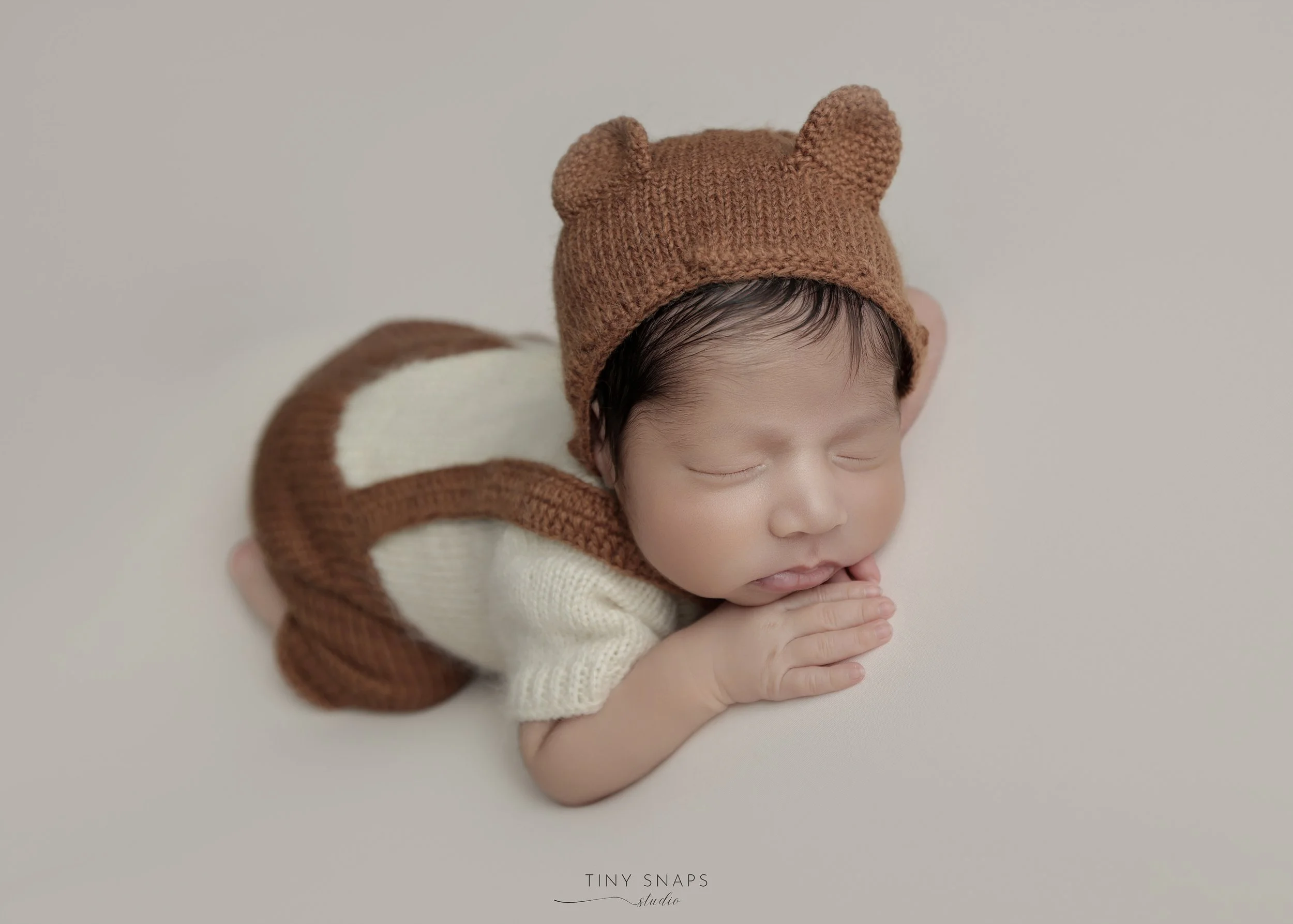 Close-up of a sleeping baby wearing a brown bear hat with ears, a cream-colored sweater, and a matching brown and cream outfit, lying on a light-colored surface with hands clasped under the chin.