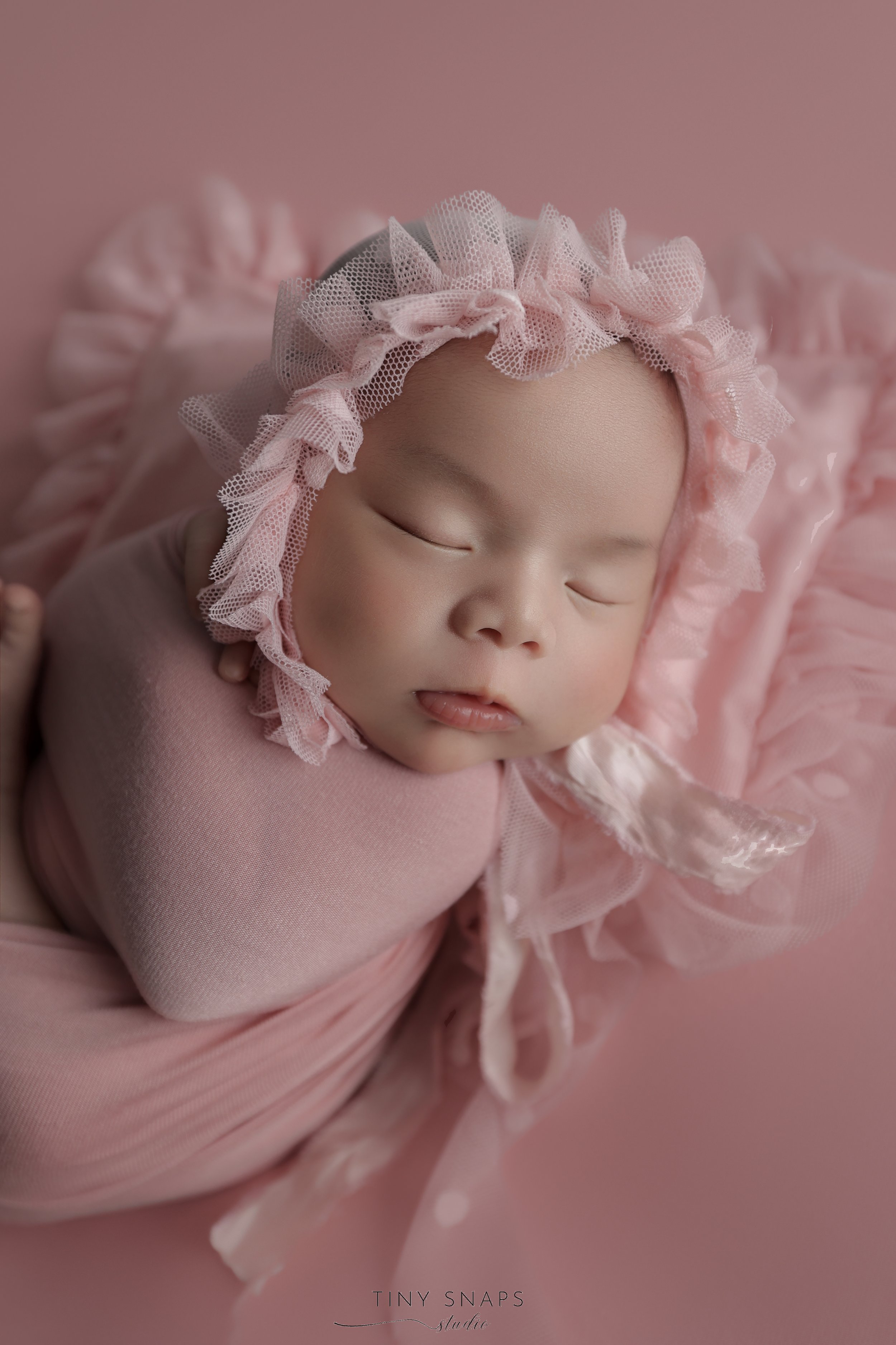 A sleeping baby wrapped in pink fabric, wearing a pink bonnet with ruffled tulle trim, on pink background.