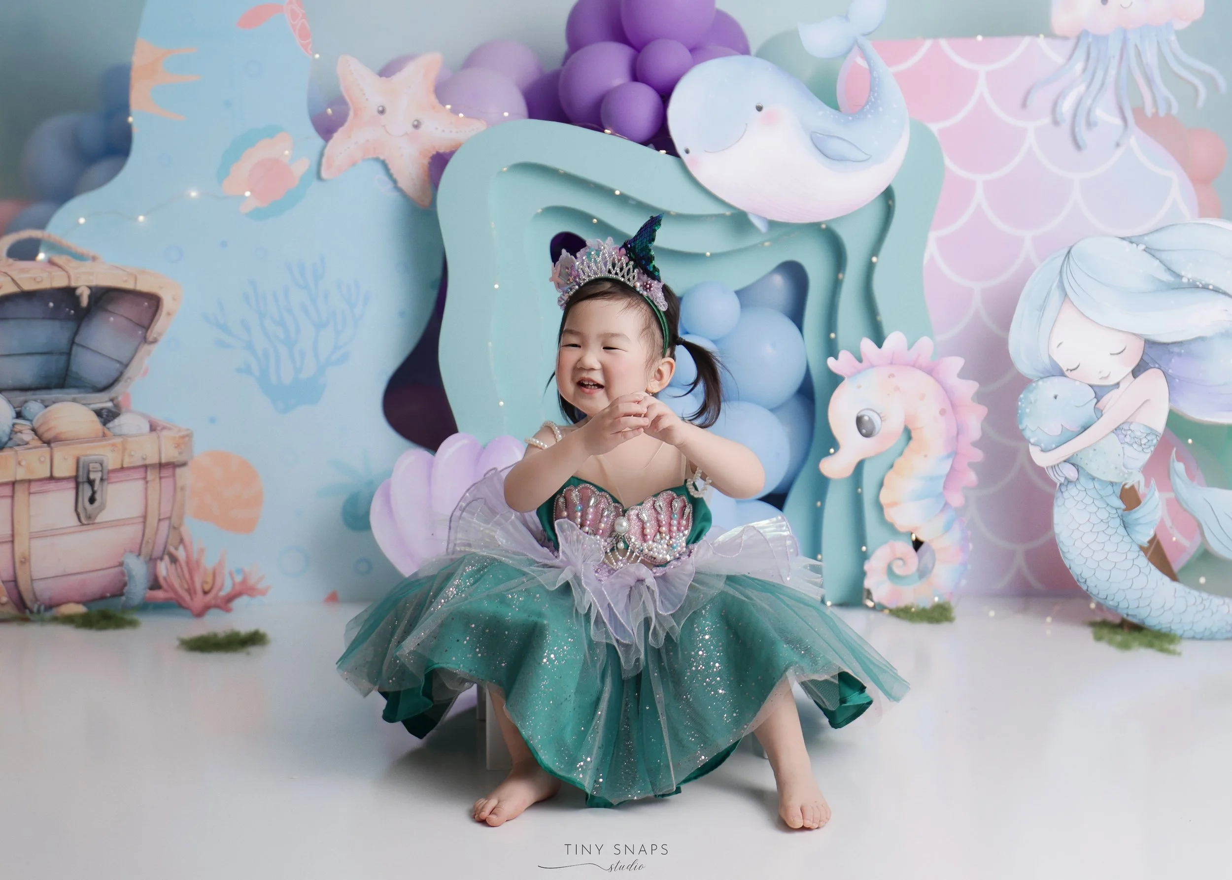 Young girl in a mermaid costume, sitting barefoot on the floor with a big smile. She is wearing a tiara and a sparkling green and purple dress. The background features an under-the-sea theme with cartoon sea creatures, treasure chest, and coral decor
