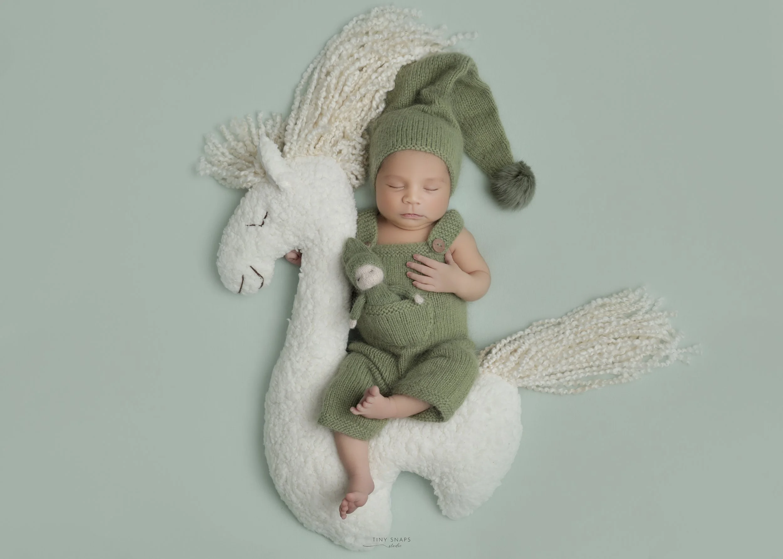 A sleeping baby in green knitted overalls and a matching hat with a pom-pom, lying on a large white plush horse with a fluffy mane and tail, on a plain mint green background.