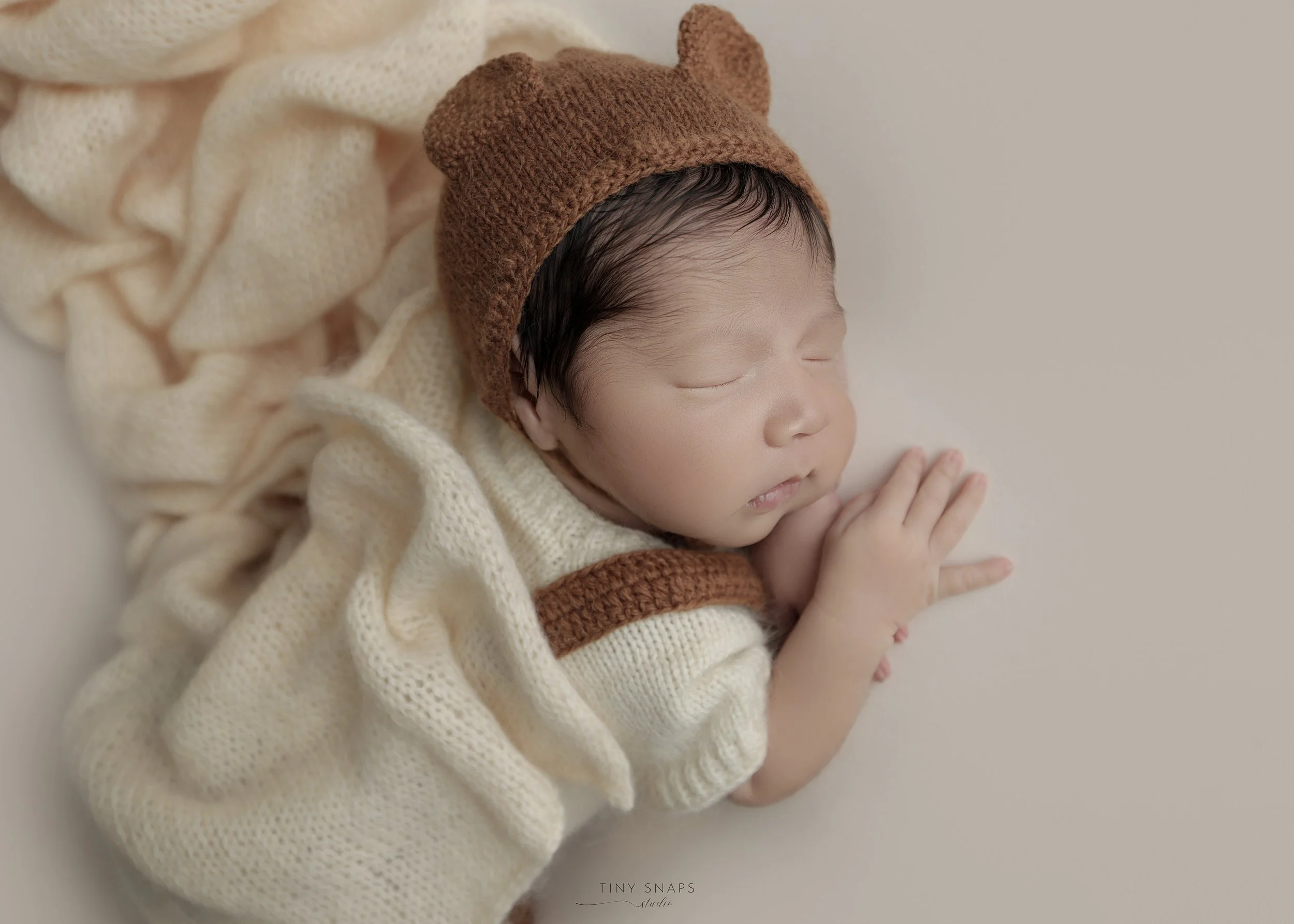 A sleeping newborn baby wearing a brown knitted hat with bear ears, cream-colored knitted sweater, and lying on a cream-colored blanket.
