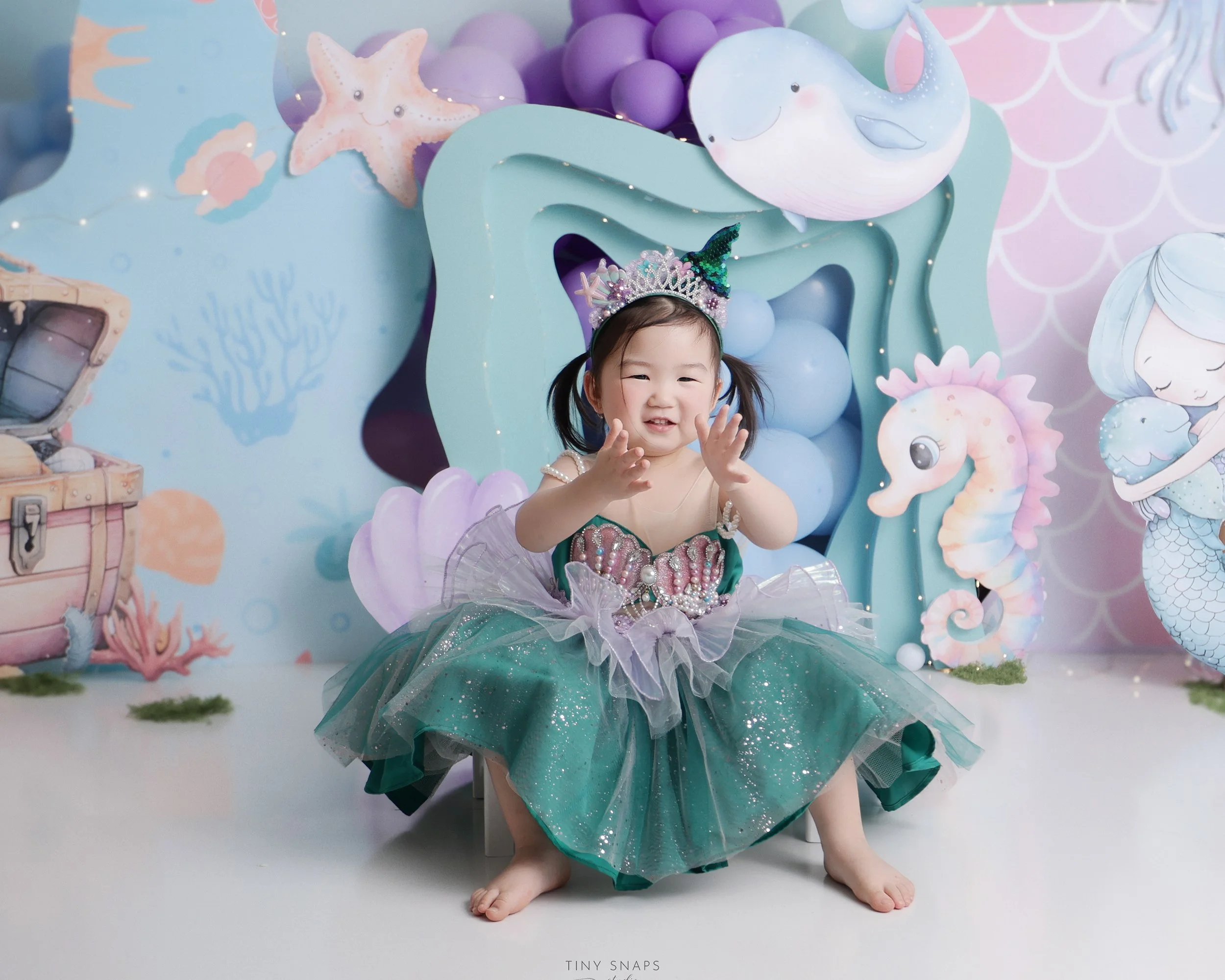 A young girl dressed as a princess in a teal and purple tutu dress with pearls and tulle, wearing a crown headband with an added unicorn horn, sitting on a small stool. She is smiling and clapping her hands, surrounded by an underwater-themed backgro