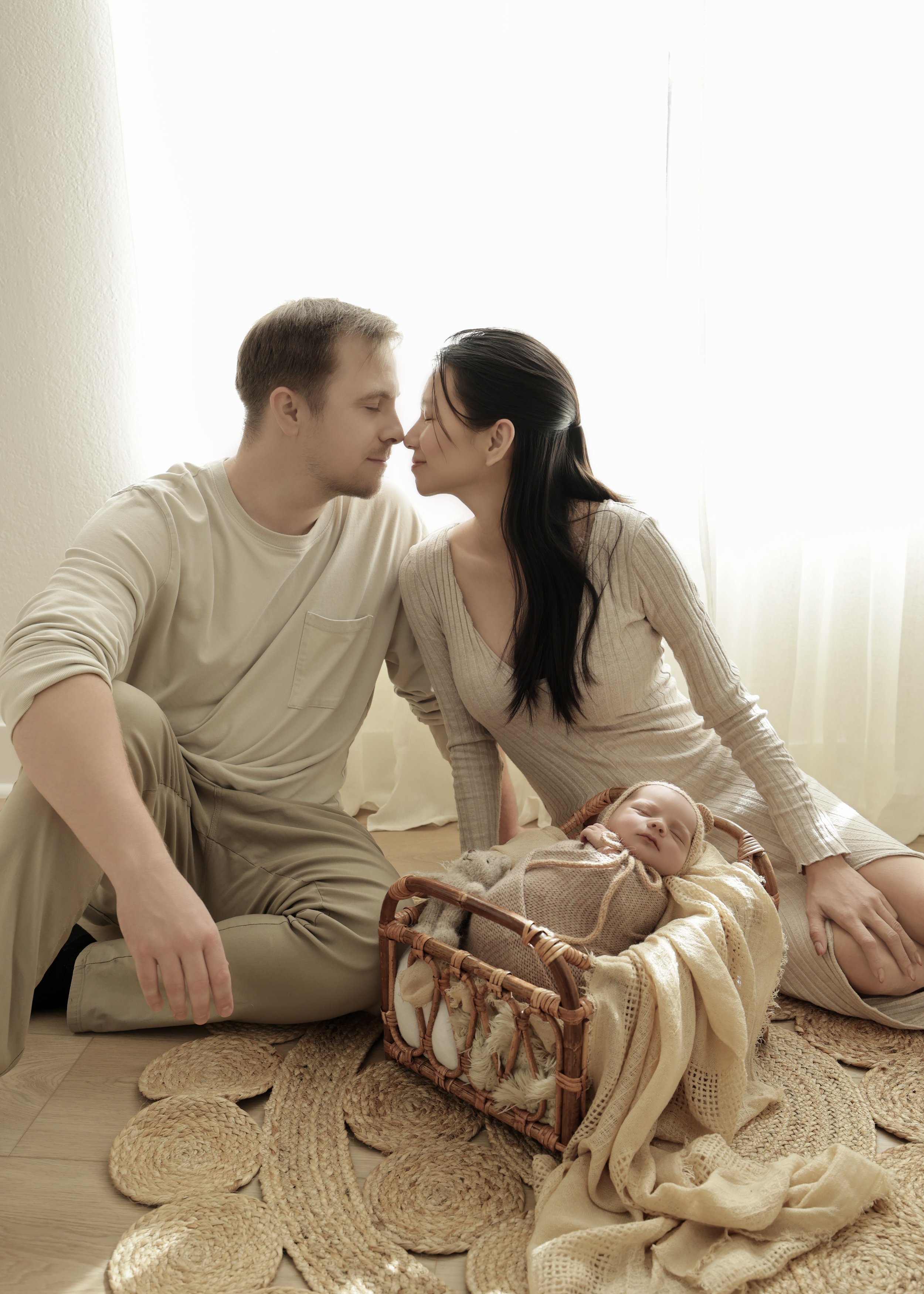 A couple sitting on the floor with their noses touching, facing each other affectionately, with a sleeping baby in a basket nearby, in a cozy, softly lit room.