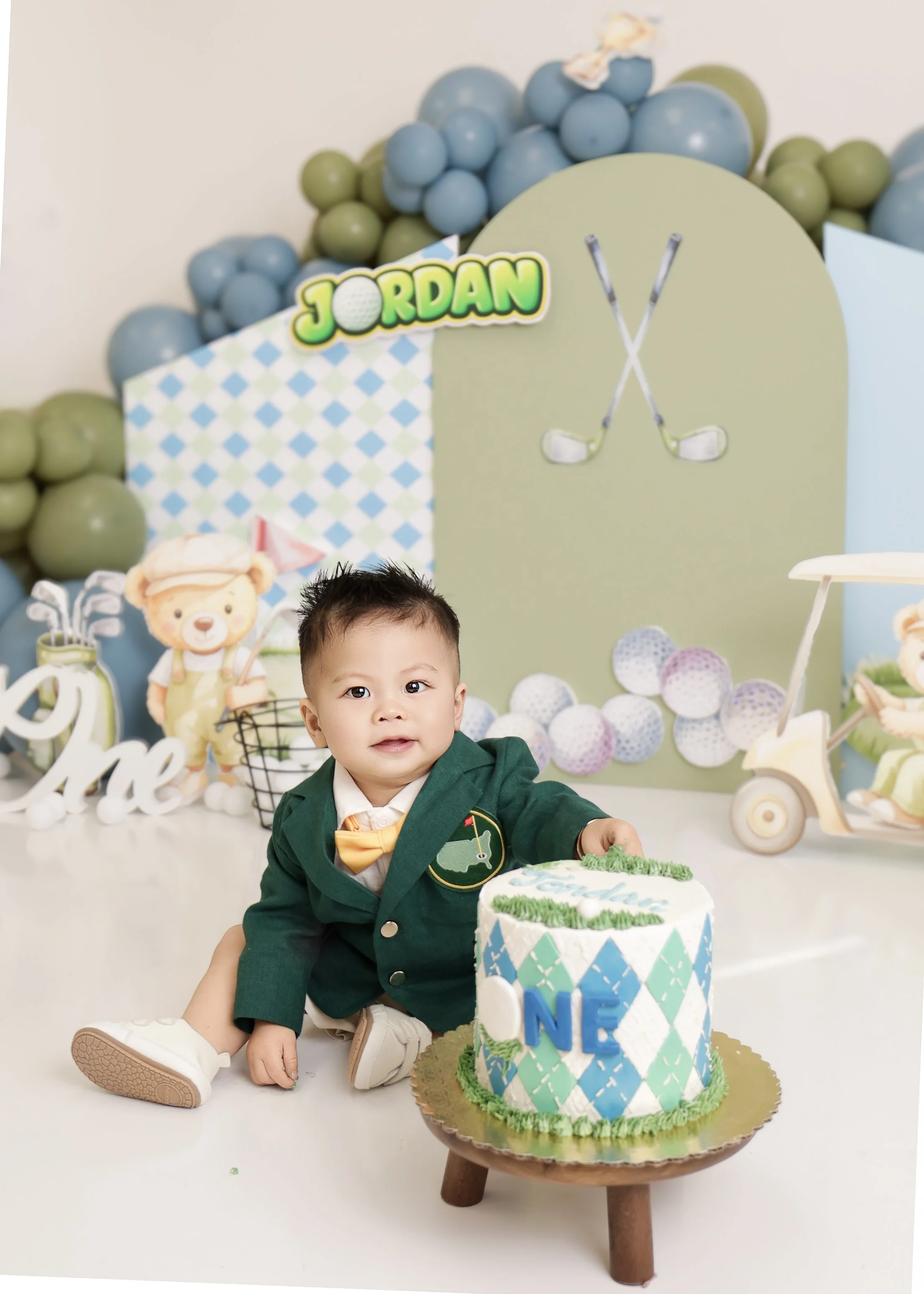 Young boy sitting next to a birthday cake with a golf theme, celebrating his first birthday, in front of a decorated backdrop with balloons and stuffed animal toys.