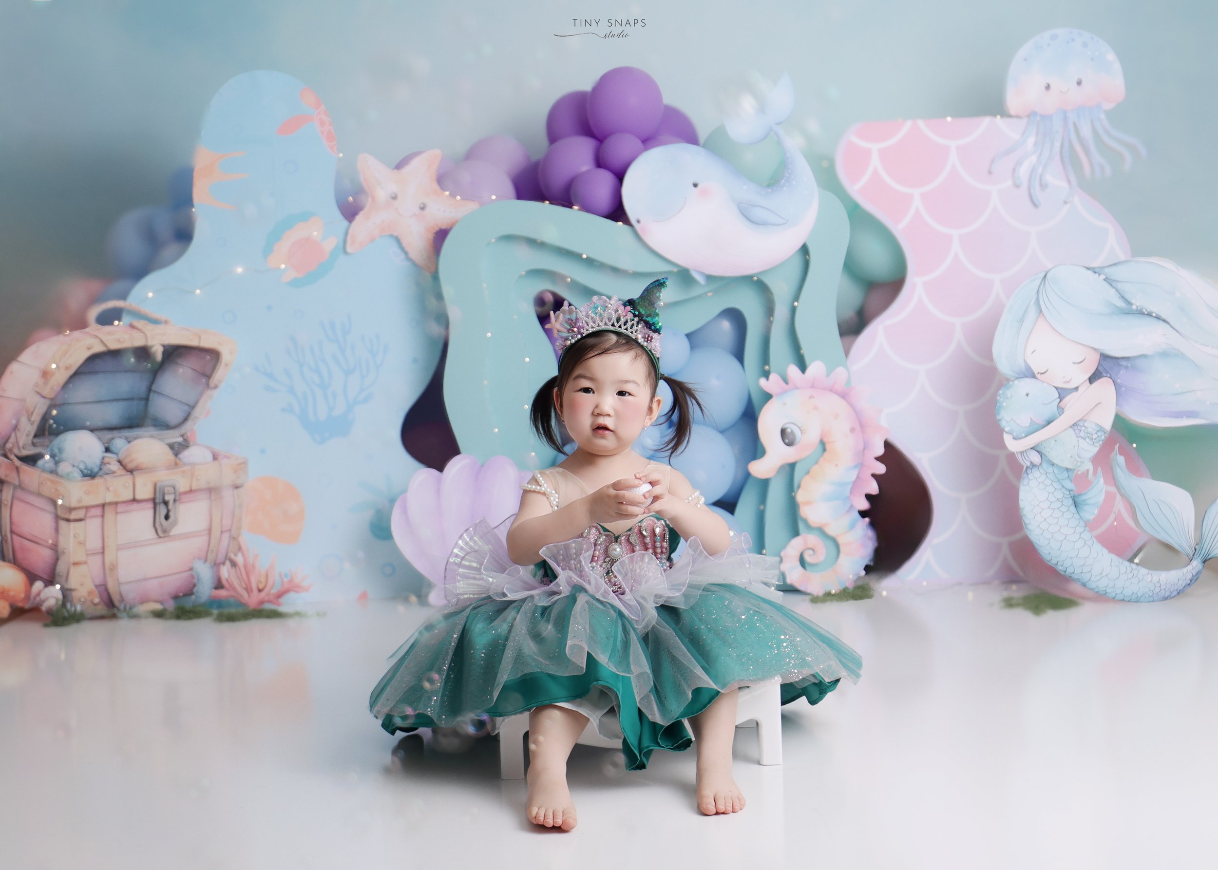 Young girl dressed as a mermaid sitting on a small white chair, wearing a tiara with a unicorn horn, with an underwater-themed backdrop featuring a treasure chest, sea creatures, and mermaids.