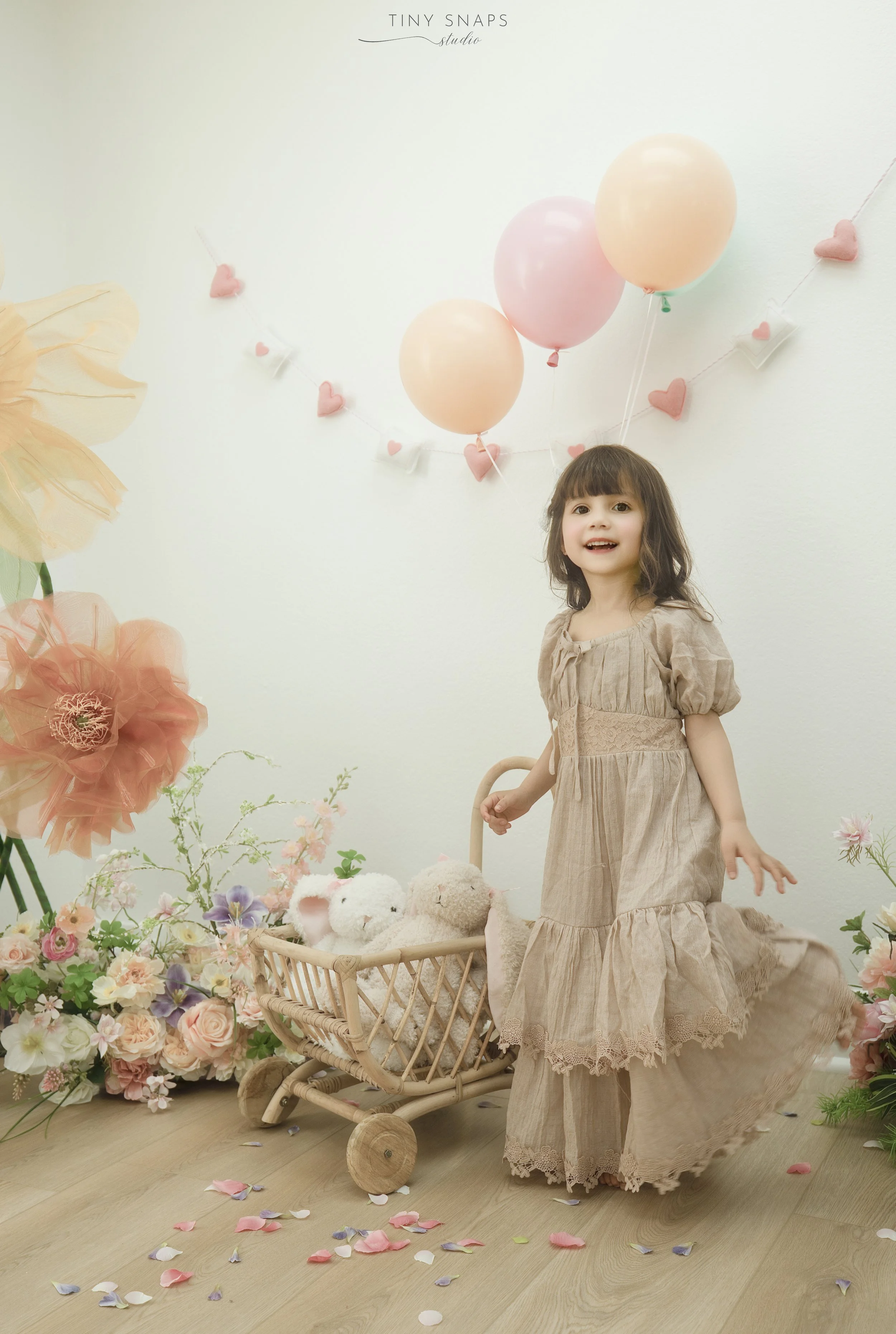 A young girl in a beige dress standing next to a small wooden toy cart filled with stuffed animals, surrounded by colorful flowers and pastel balloons, with pink and purple flower petals scattered on the floor, and decorative hearts and balloons hang