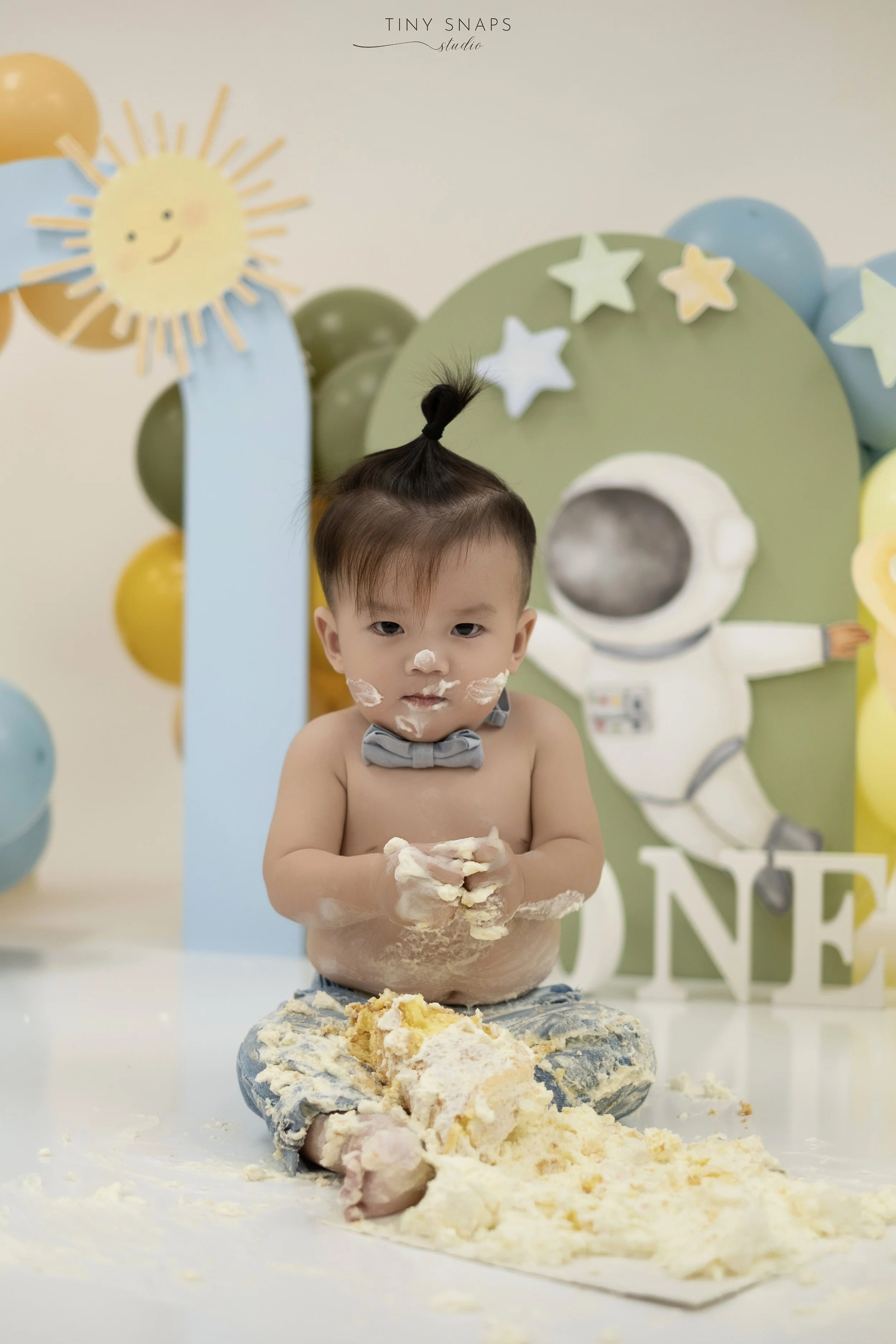 A young child with messy cake and frosting on his face and hands, sitting on the floor in front of a space-themed birthday backdrop with balloons, a smiling sun, stars, and an astronaut figure. The child is shirtless, wearing a gray bow tie, and appears to be celebrating his first birthday.