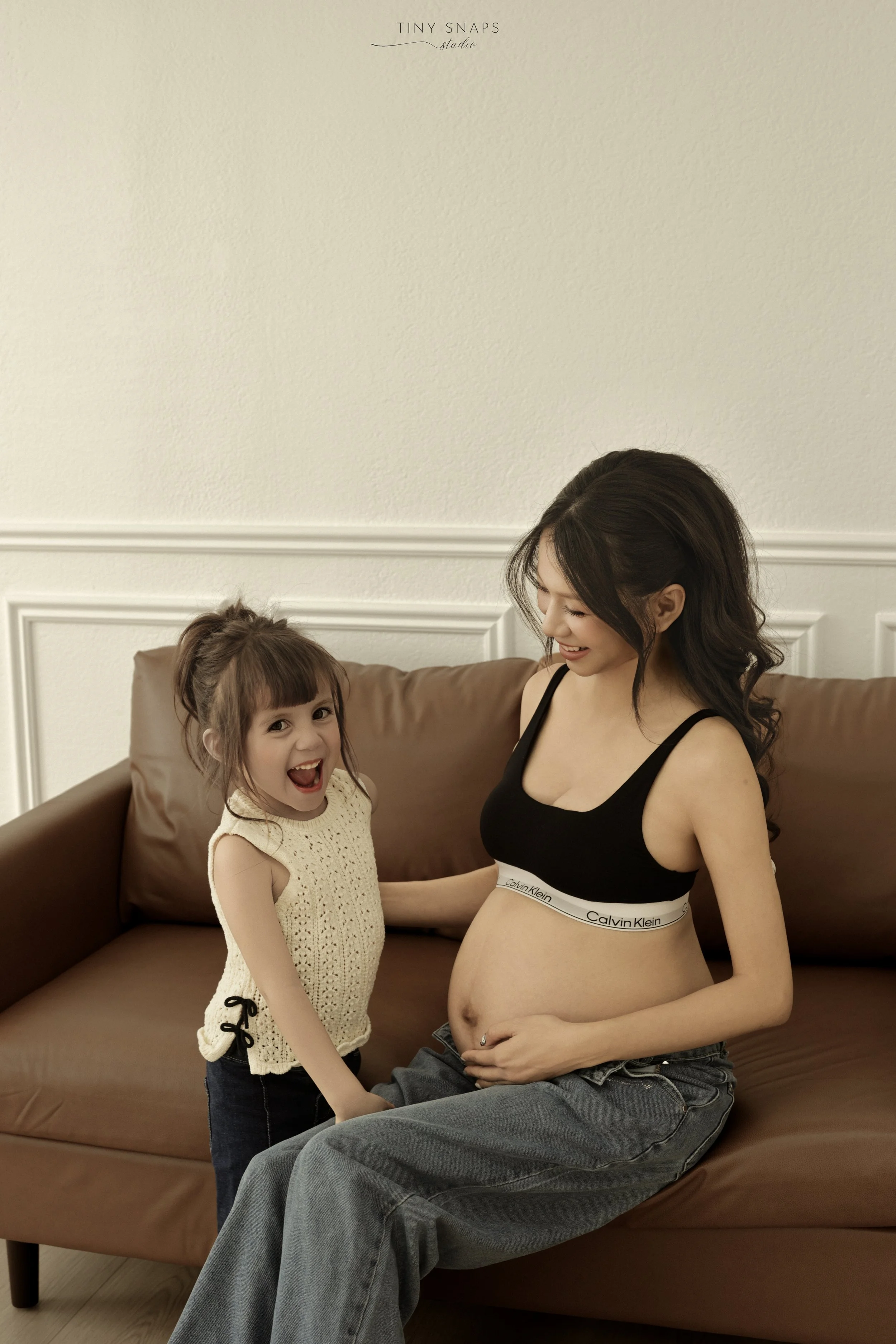 A pregnant woman and a young girl sitting on a brown sofa, smiling and enjoying each other's company in a cozy living room.