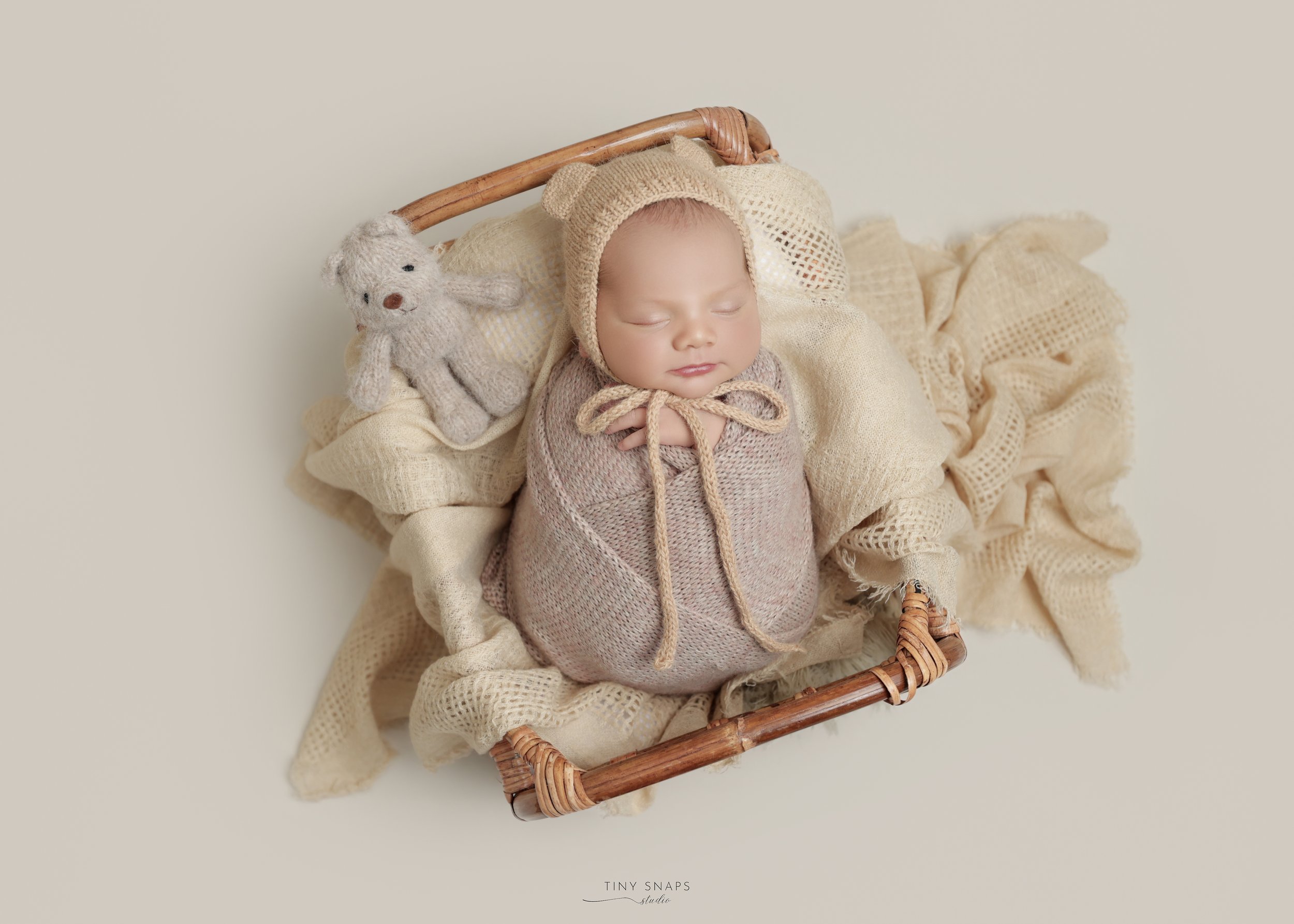 A sleeping baby dressed in a knitted outfit with a matching hat, lying in a basket lined with cozy blankets, with a small knitted teddy bear nearby.