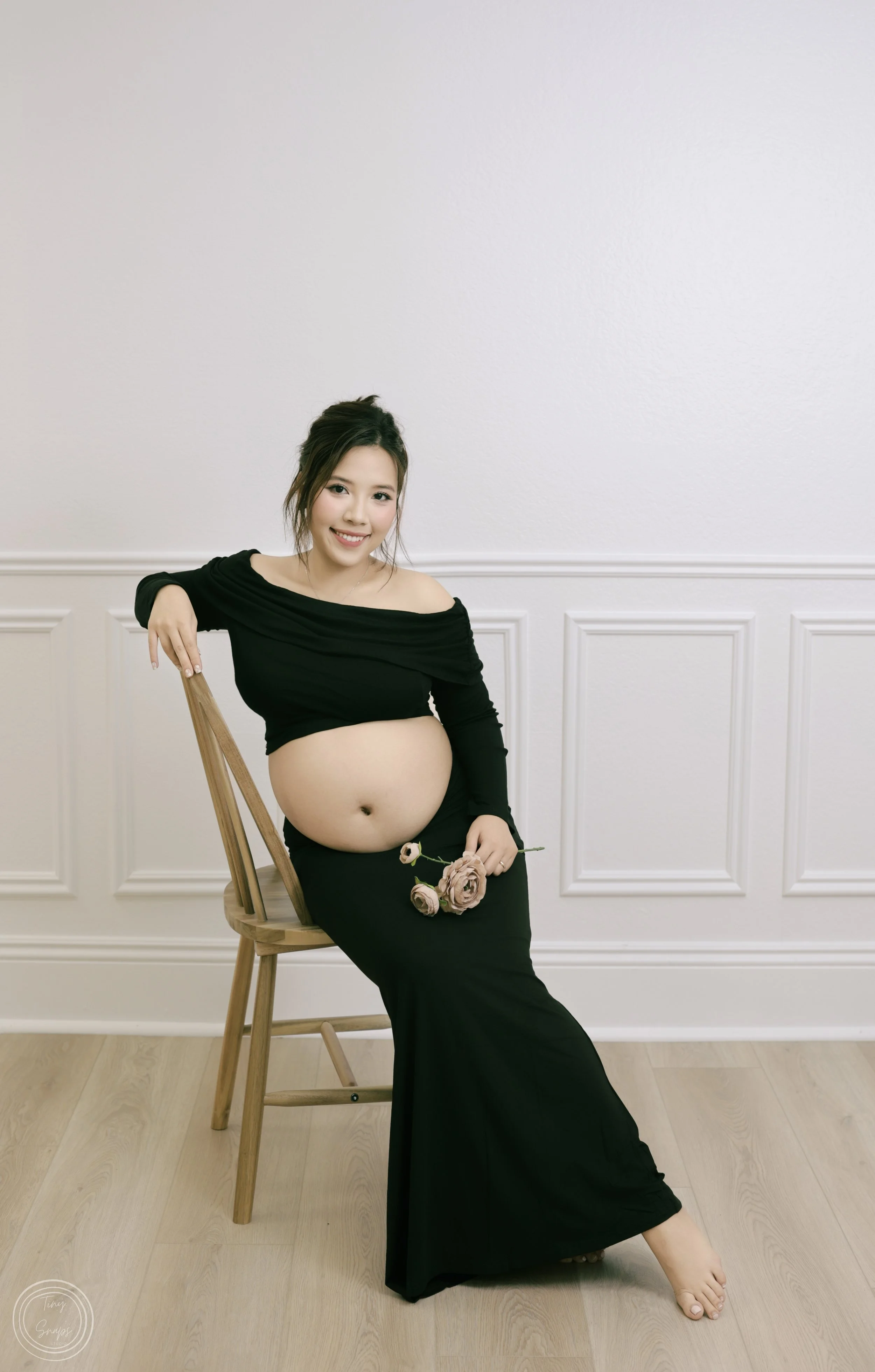 A pregnant woman in a black off-the-shoulder dress sitting on a wooden chair, holding a bunch of pink flowers, smiling against a white wall with decorative molding.