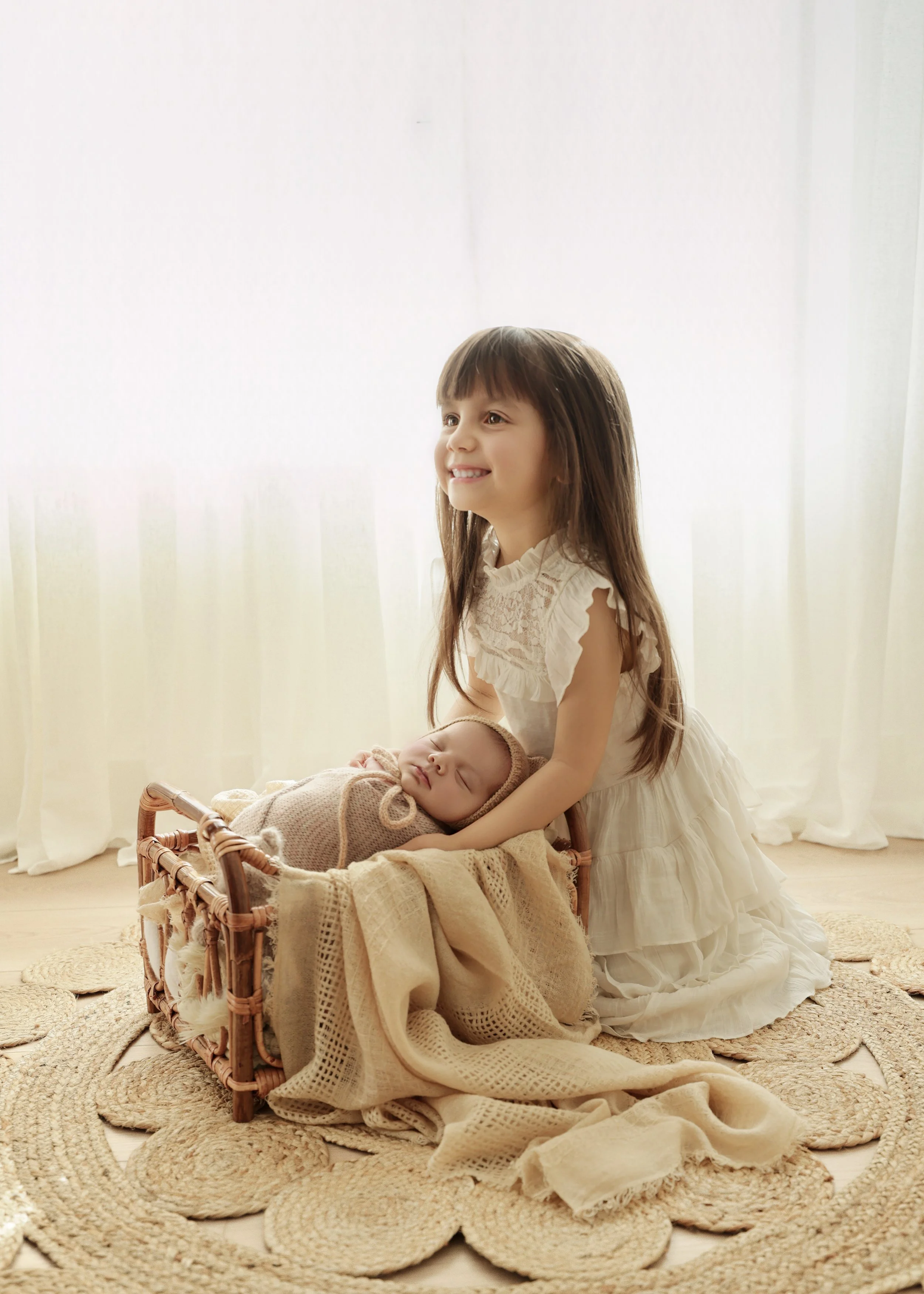A young girl in a white dress kneeling on a woven rug, smiling, with a sleeping baby in a basket lined with a beige knitted blanket, in front of sheer white curtains.