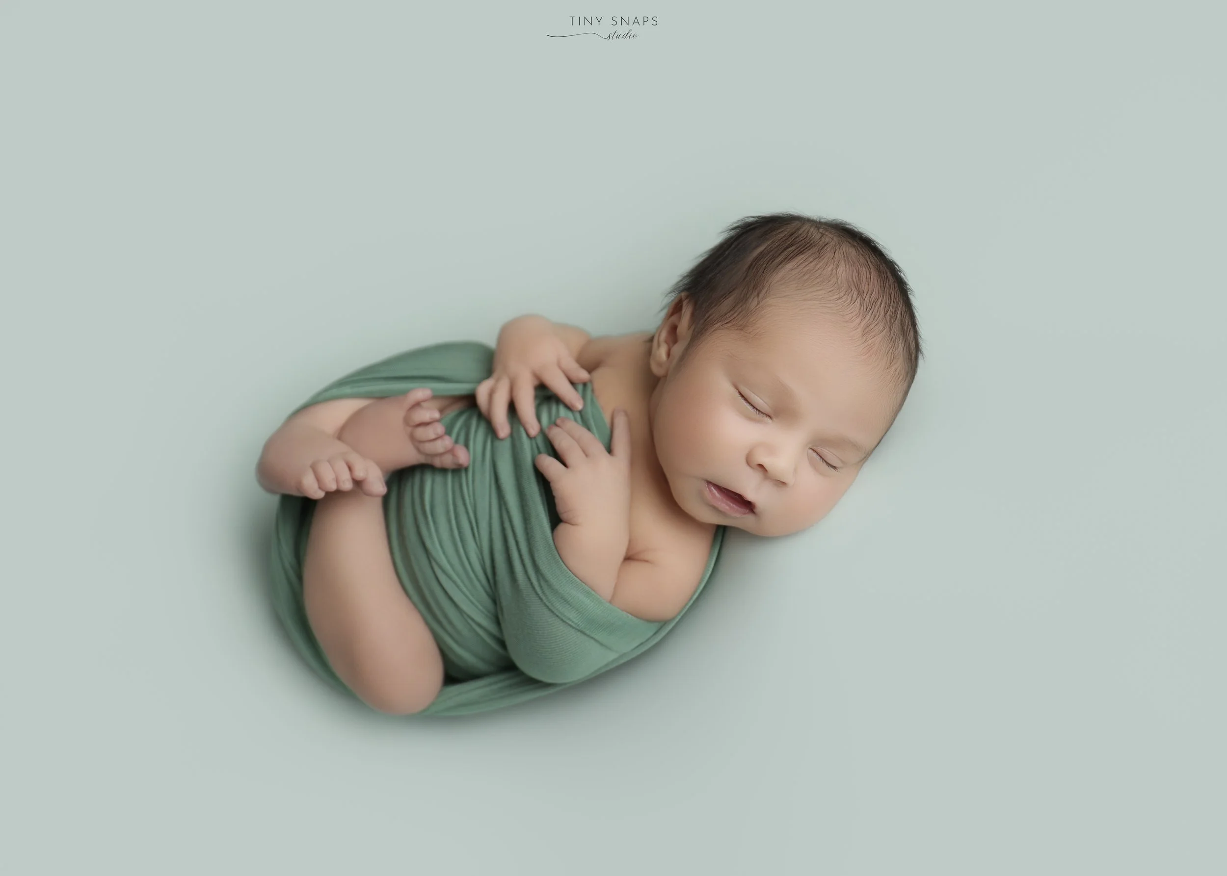 A sleeping newborn baby lying on a light blue background, wearing a green onesie, with hands near the face and eyes closed.