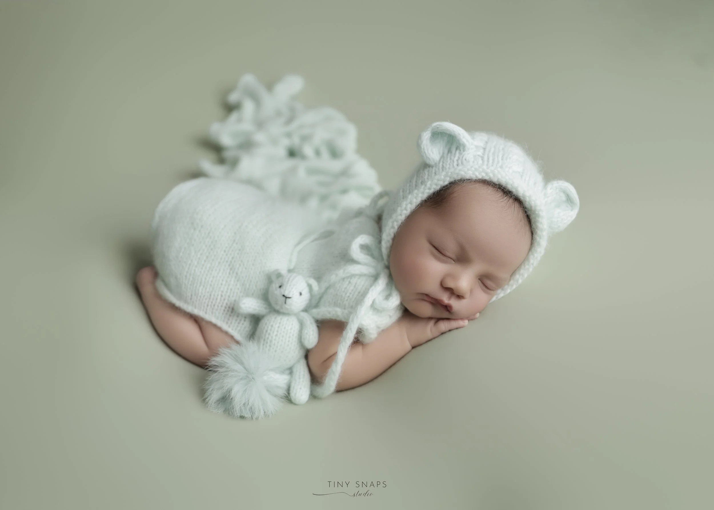 Sleeping baby wearing a white knit hat with bear ears, laying on a soft surface, with a small knit teddy bear. The baby has a peaceful expression, and the background is a plain light color.