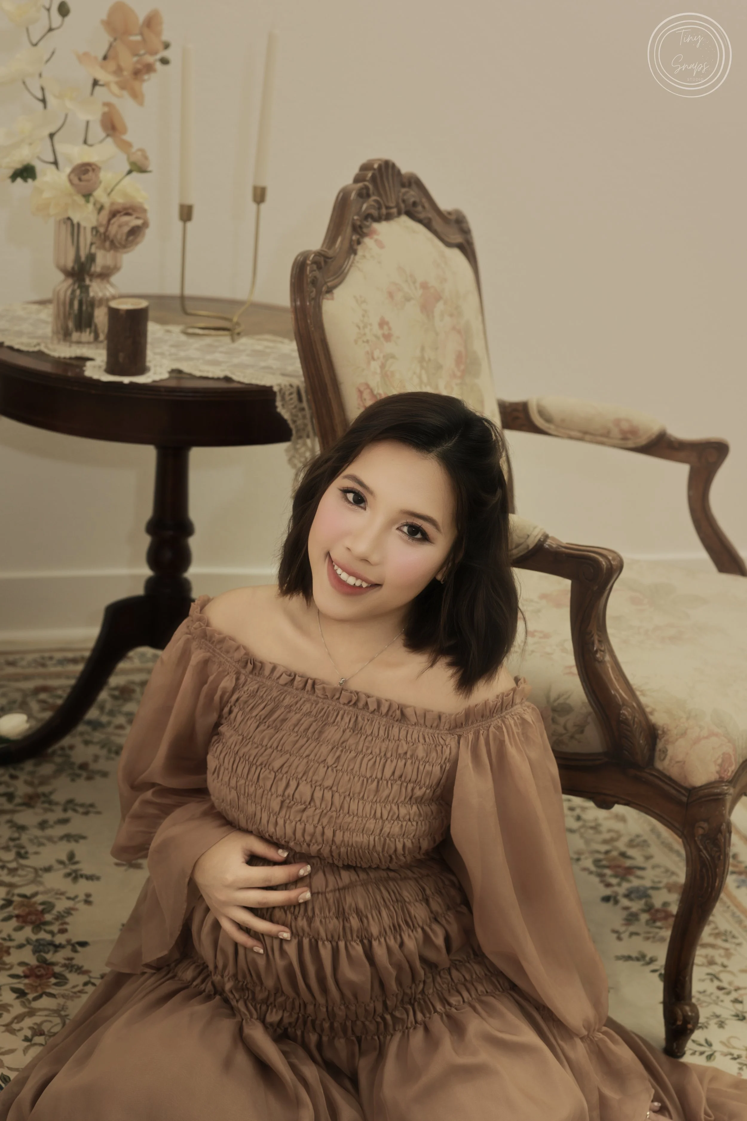 A young woman with shoulder-length dark hair, smiling, sitting on the floor in front of a vintage-style armchair with a floral pattern. She wears a light brown off-shoulder dress. Behind her is a wooden table with a lace runner, a flower vase with wh