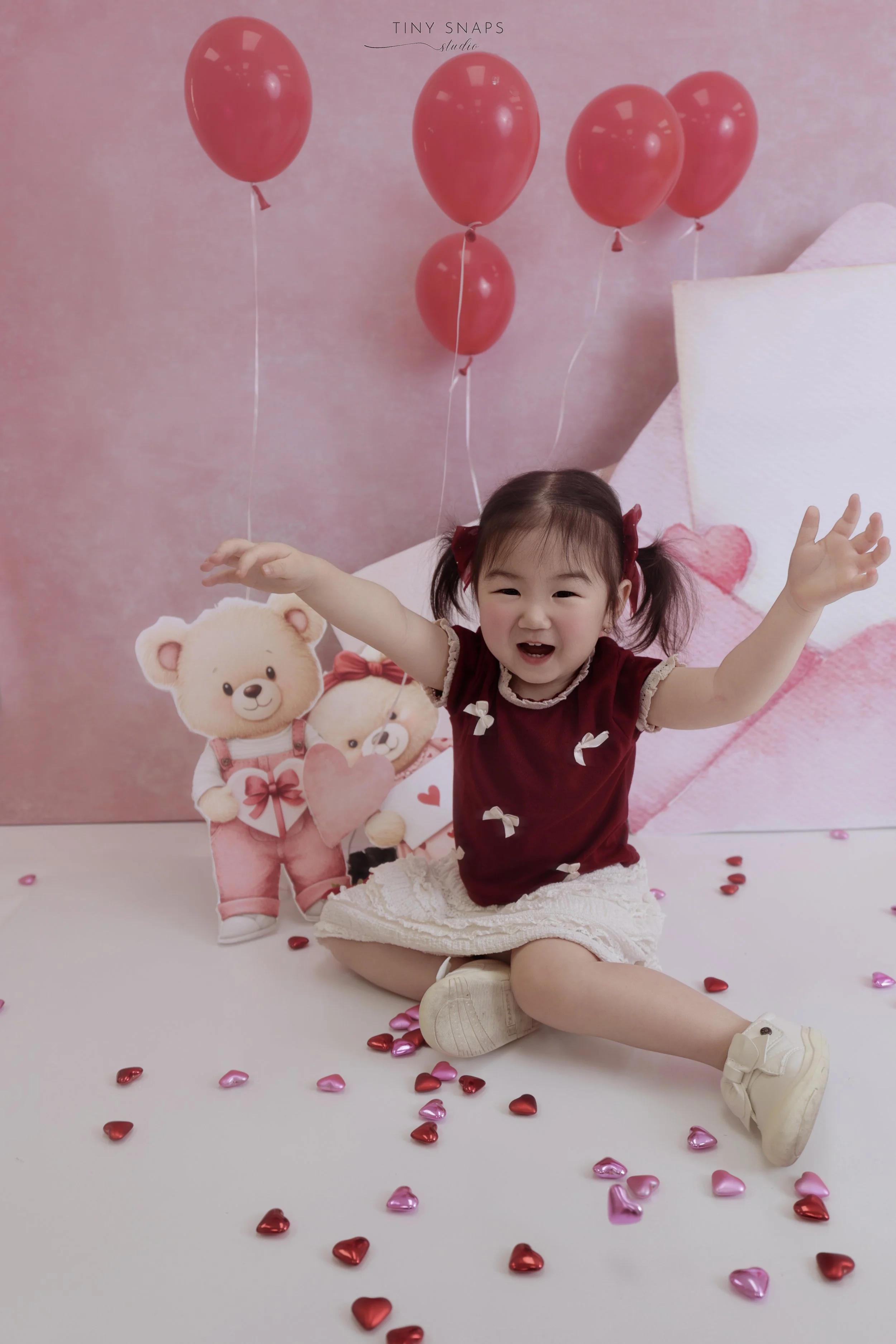 A young girl in a red top and white skirt sitting on a white floor, smiling and raising her arms, surrounded by pink and red hearts on the floor. Behind her, a pink background features four pink balloons floating above cartoon teddy bear and bear hol