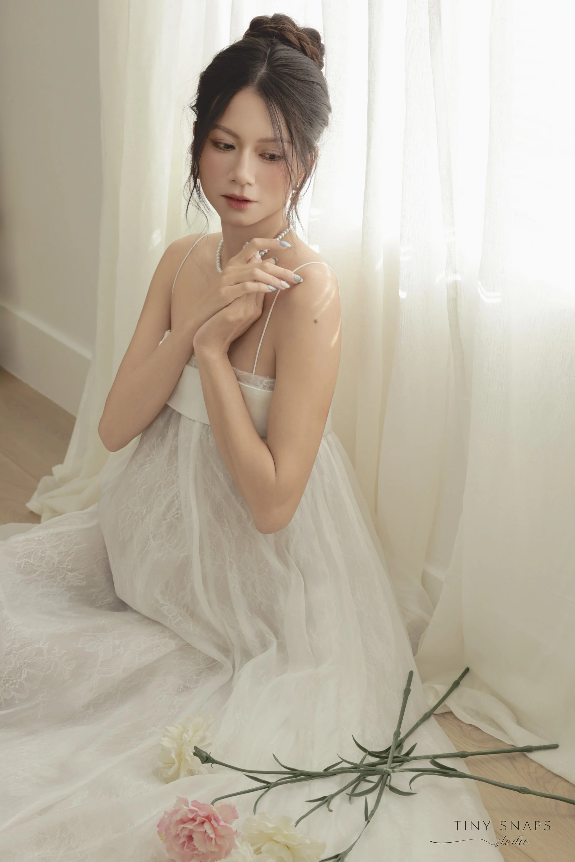 A young woman with dark hair styled in an updo, sitting on a wooden floor near a sheer white curtain, wearing a white lace dress and jewelry, holding her hands close to her chest, with pink and white carnations lying nearby.