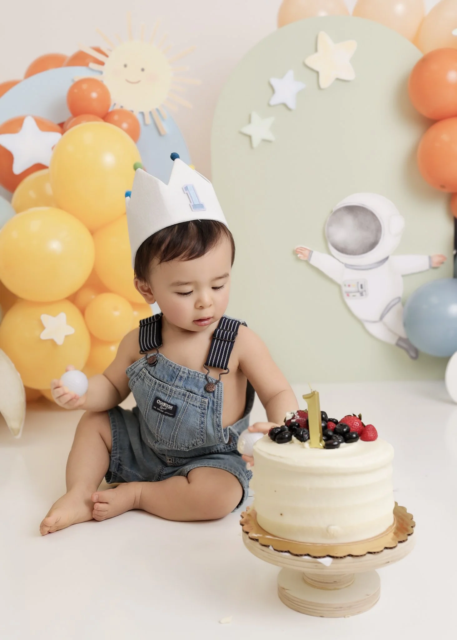 A young child wearing a birthday crown and denim overalls sitting on the floor next to a birthday cake topped with berries and a gold number 1 candle, with colorful balloons and space-themed decorations in the background.
