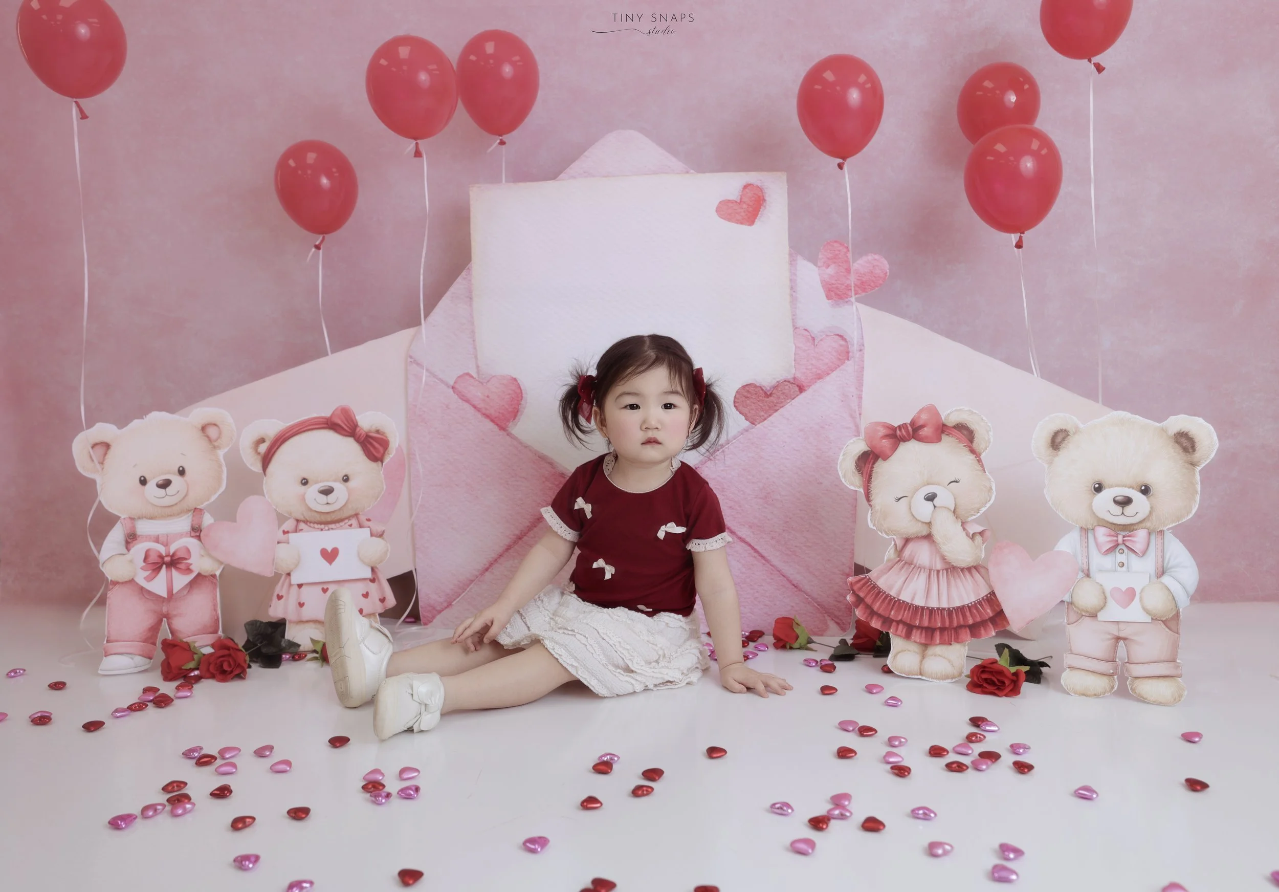 A young girl sitting on the floor surrounded by Valentine's Day decorations and stuffed teddy bear cutouts, with pink and red balloons and heart-shaped confetti in a pink-themed setting.