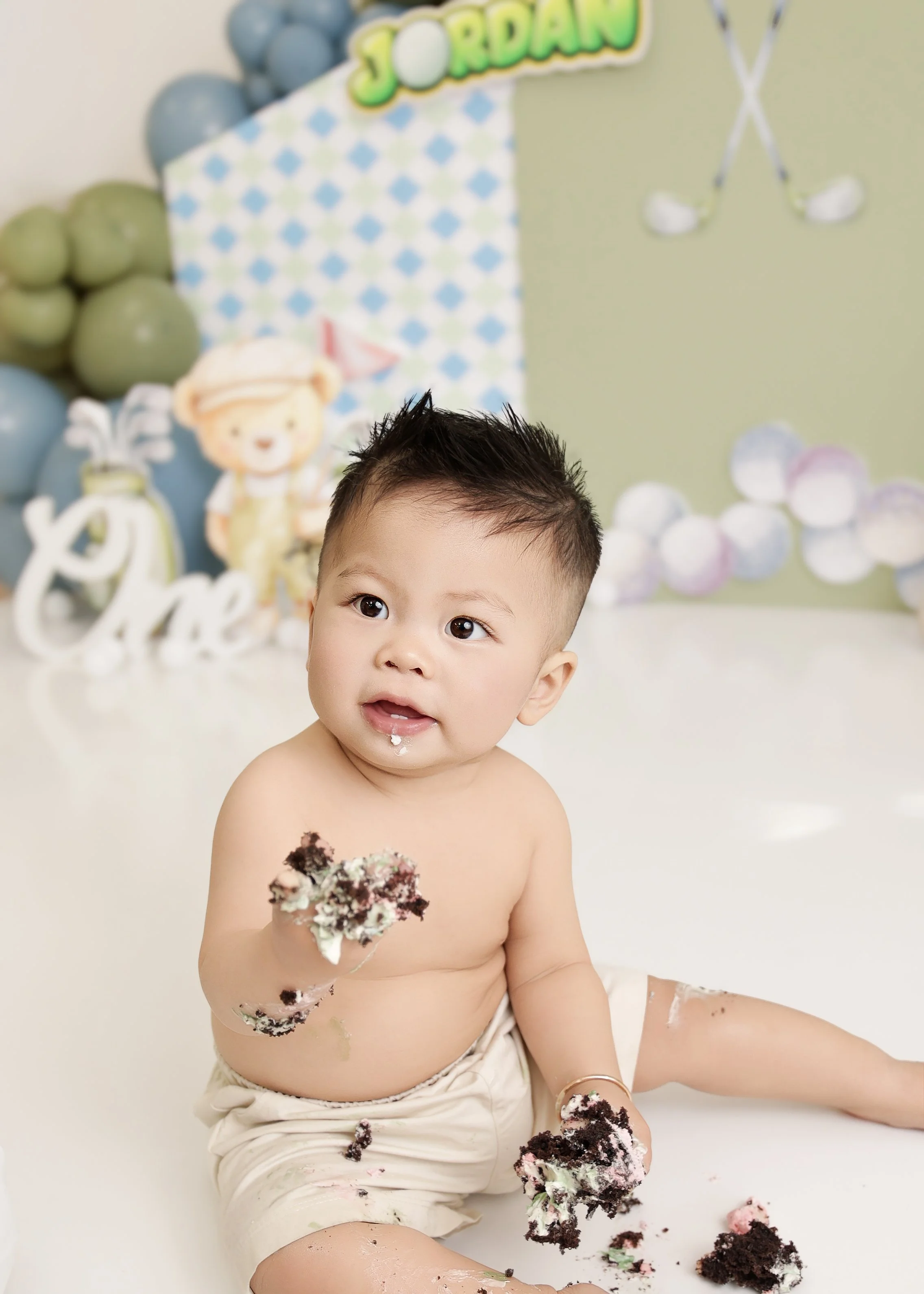Young toddler boy sitting on the floor with cake, wearing only beige shorts, with cake remnants on his face and body, in front of a birthday party backdrop with balloons and decorations.