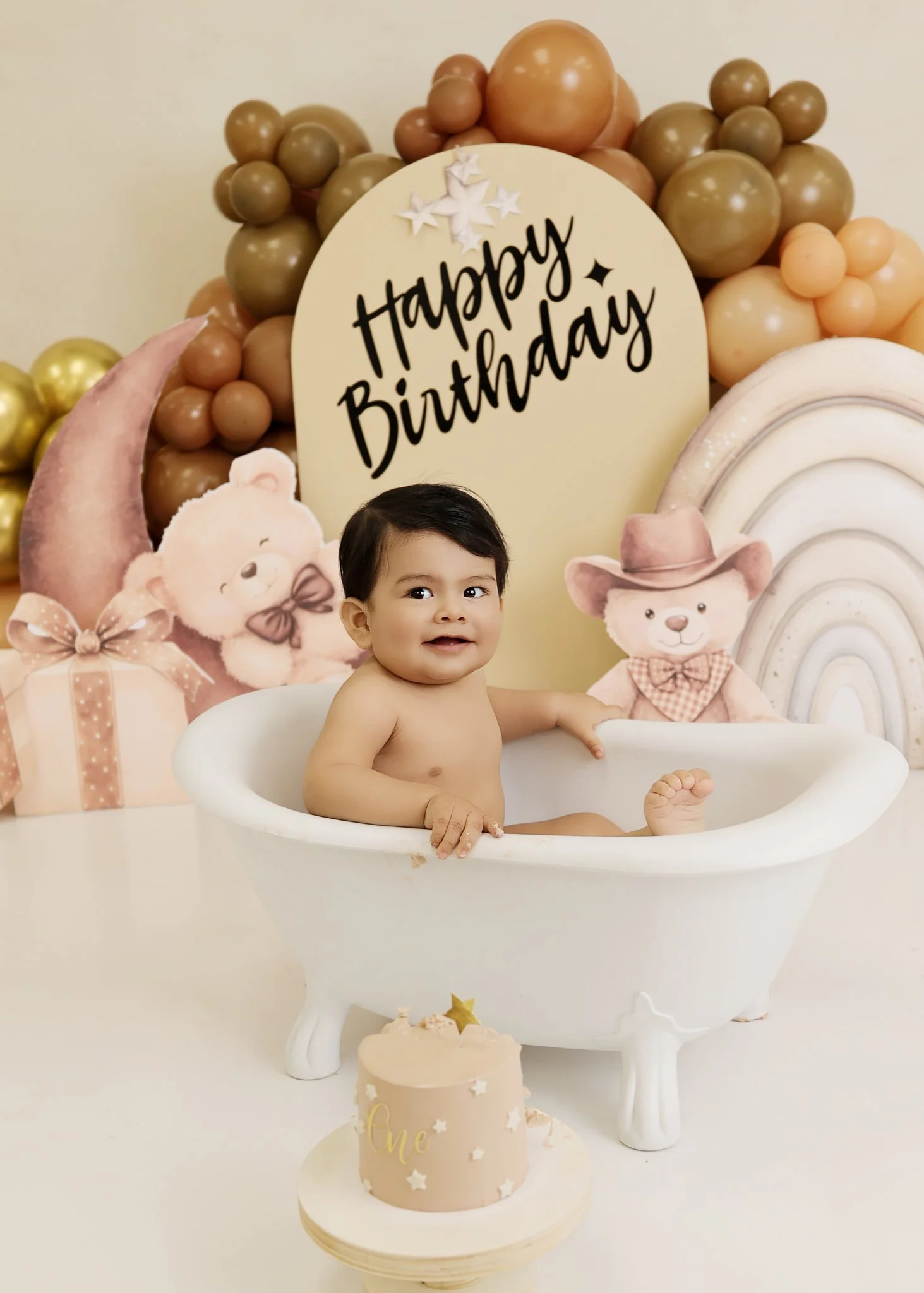 Baby sitting in a small white bathtub with a backdrop of teddy bears, balloons, and a 'Happy Birthday' sign, celebrating a first birthday with a cake in front.