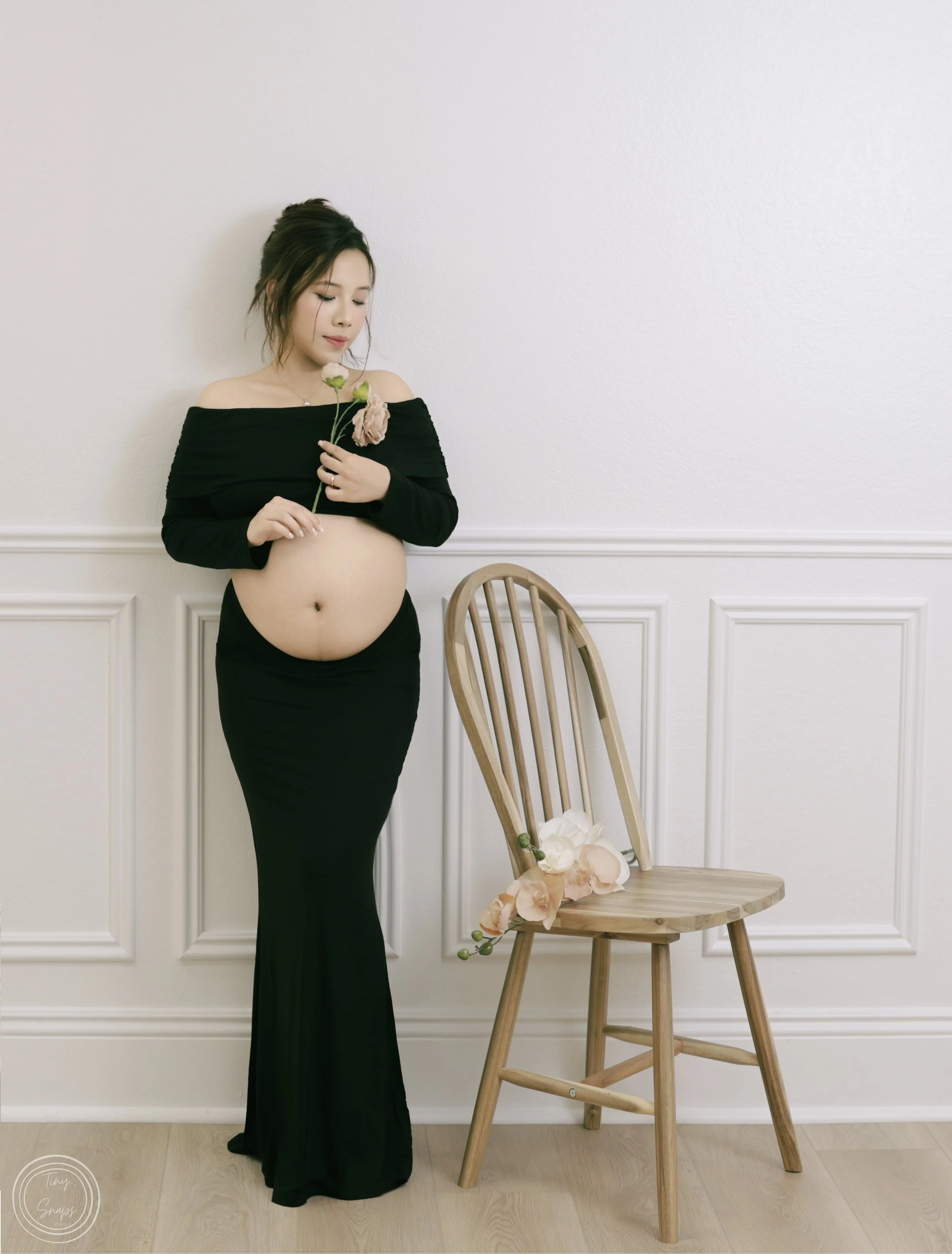 Pregnant woman in a black off-shoulder dress holding a flower, standing next to a wooden chair with flowers on it, against a white paneled wall.