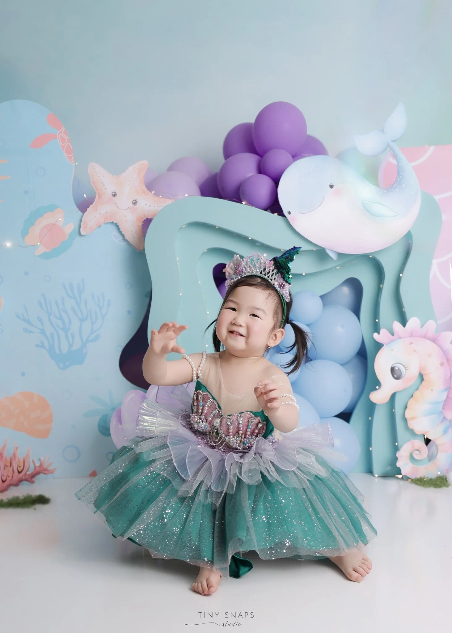 A young girl in a mermaid costume with a purple and green tulle skirt, a seashell top, and pearl accessories, smiling and waving in front of an ocean-themed backdrop with balloons, a smiling whale, starfish, seahorse, and underwater plants.