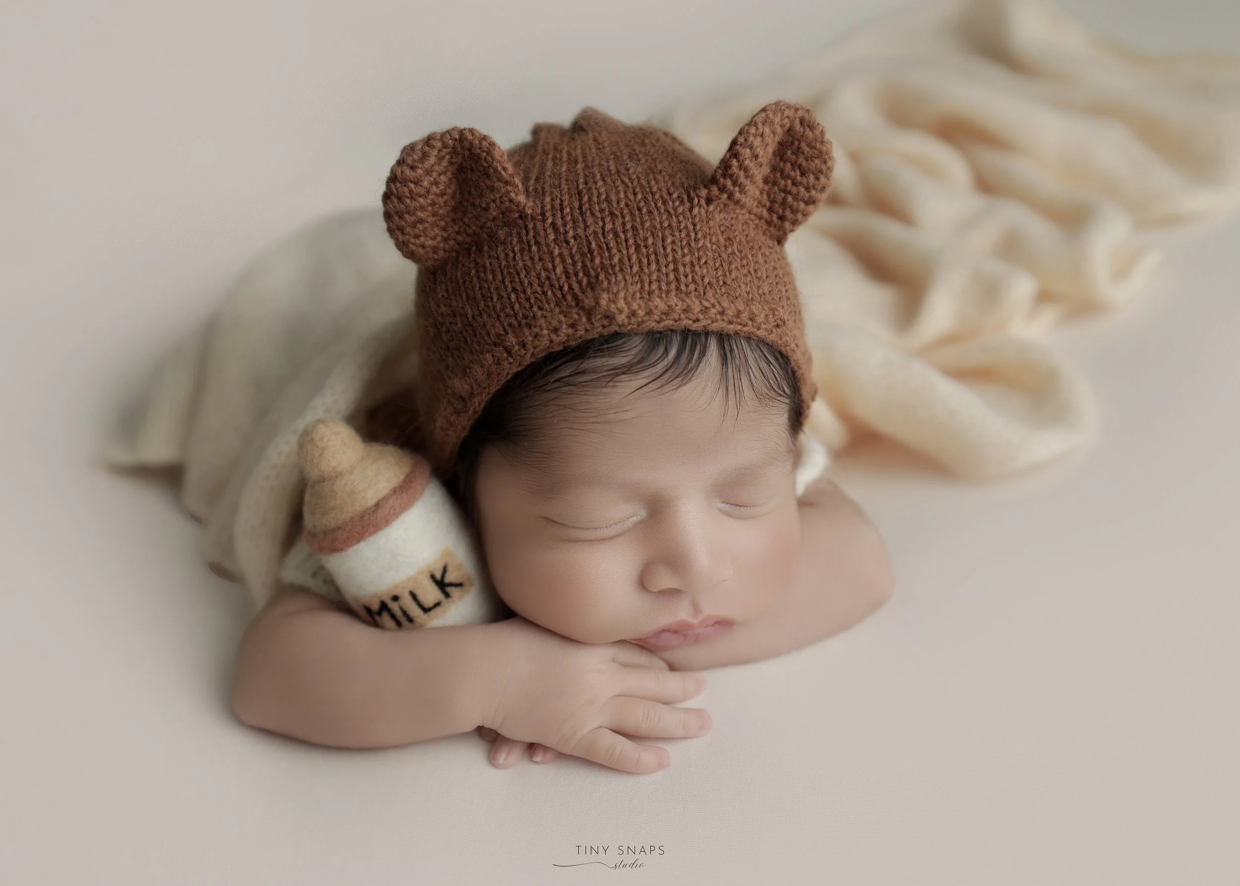 A sleeping baby wearing a brown bear hat, lying on a soft surface with a cream-colored blanket and holding a small stuffed milk bottle toy.