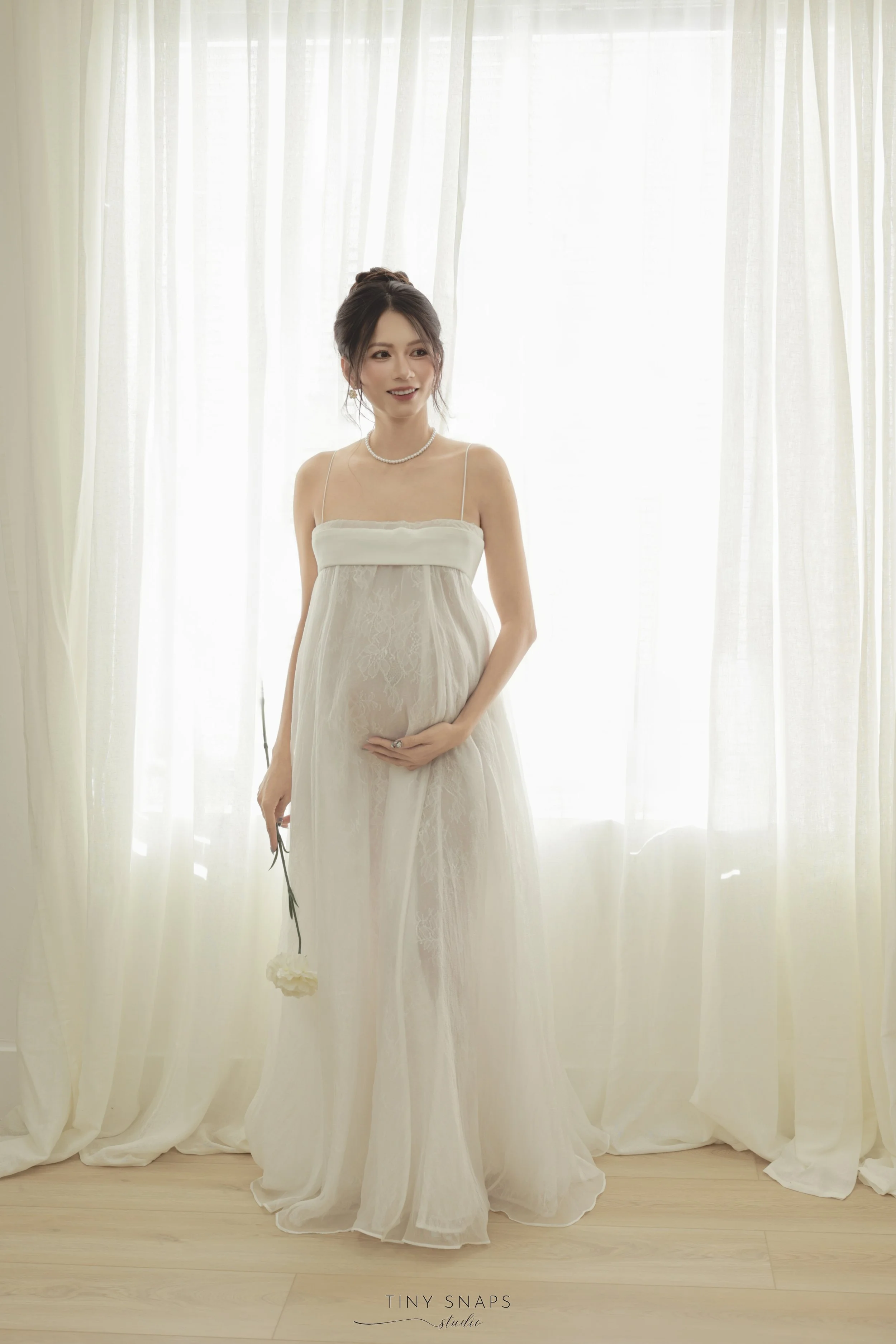 A pregnant woman wearing a white evening gown and pearl necklace standing in front of sheer curtains, holding a white flower in her right hand and resting her left hand on her belly.