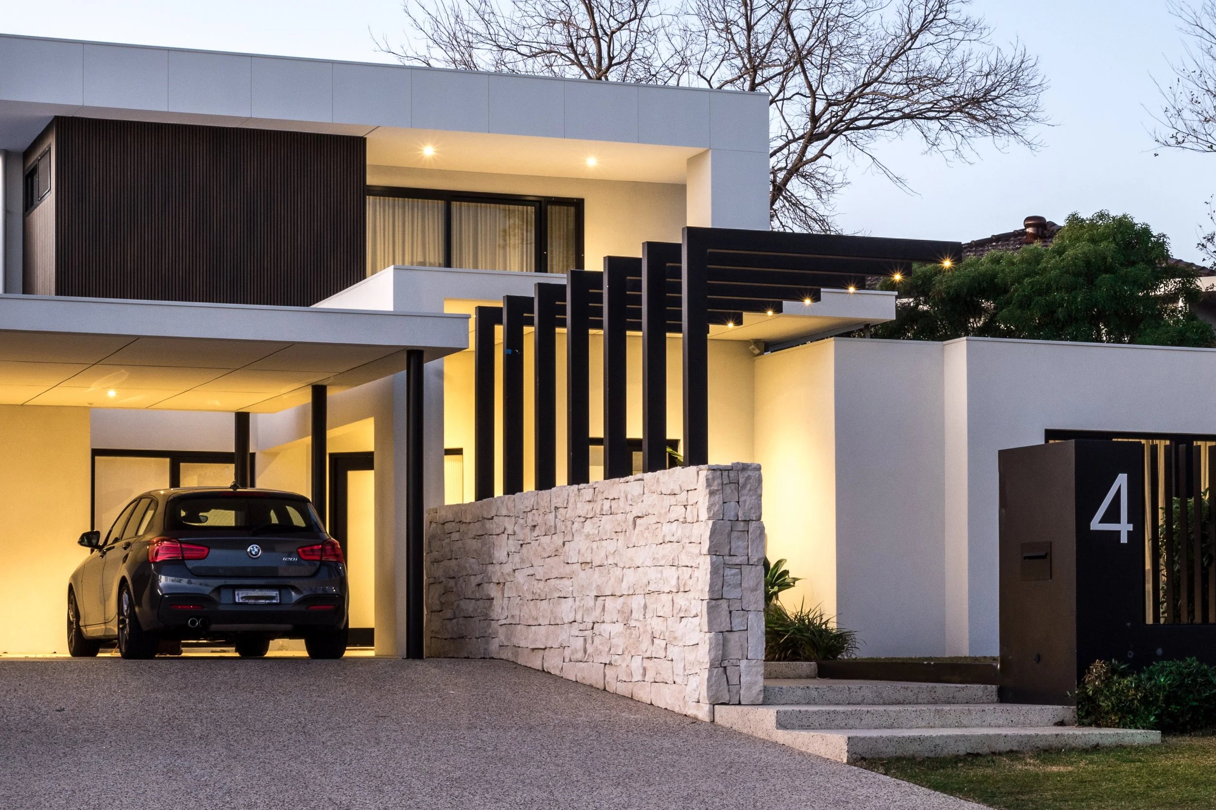 Modern two-story house with a black BMW parked in the garage, illuminated exterior lights, stone wall, black vertical slats, and the number 4 on the gate, surrounded by trees.