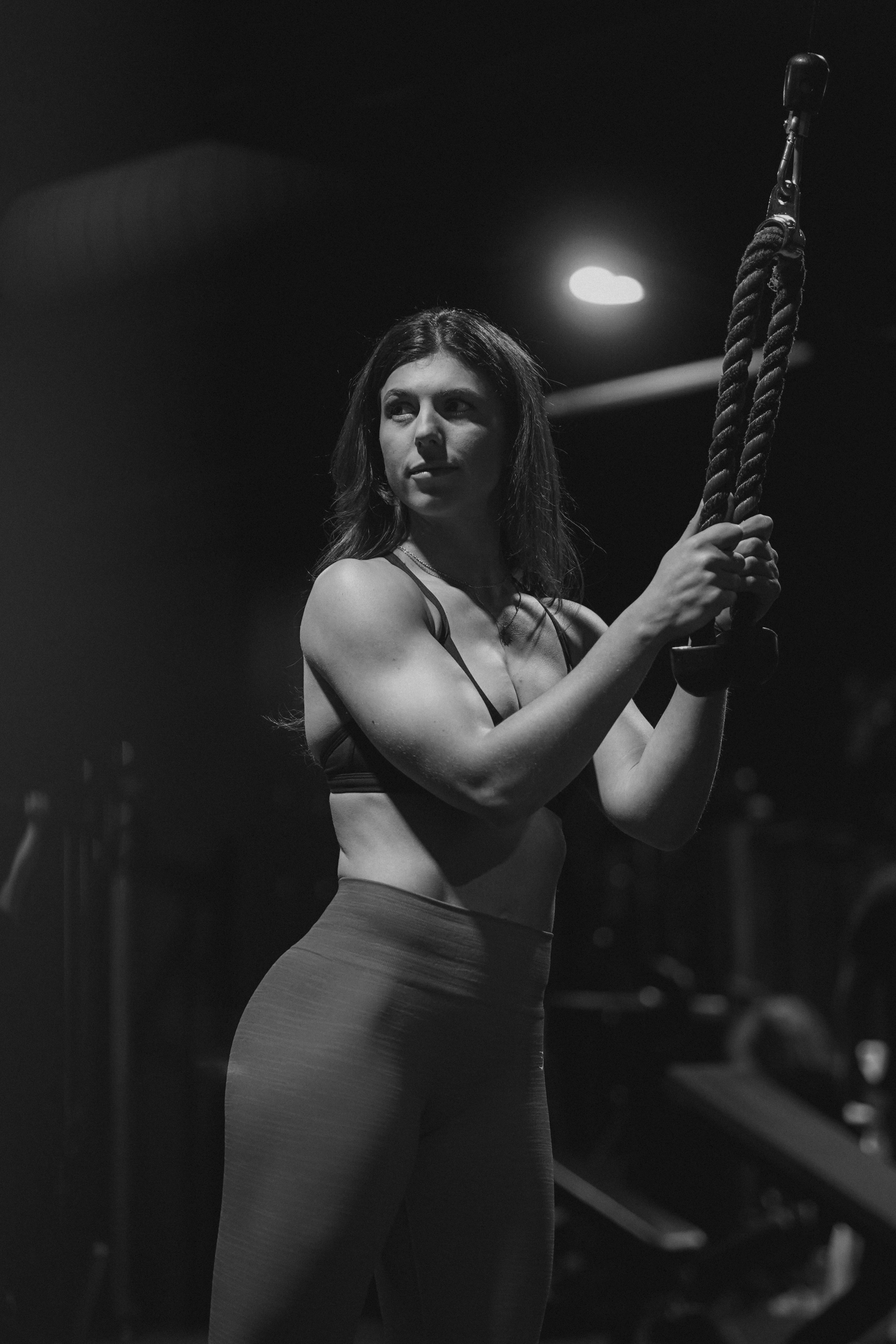 A woman in workout attire holding onto thick gym ropes in a dimly lit indoor fitness studio.