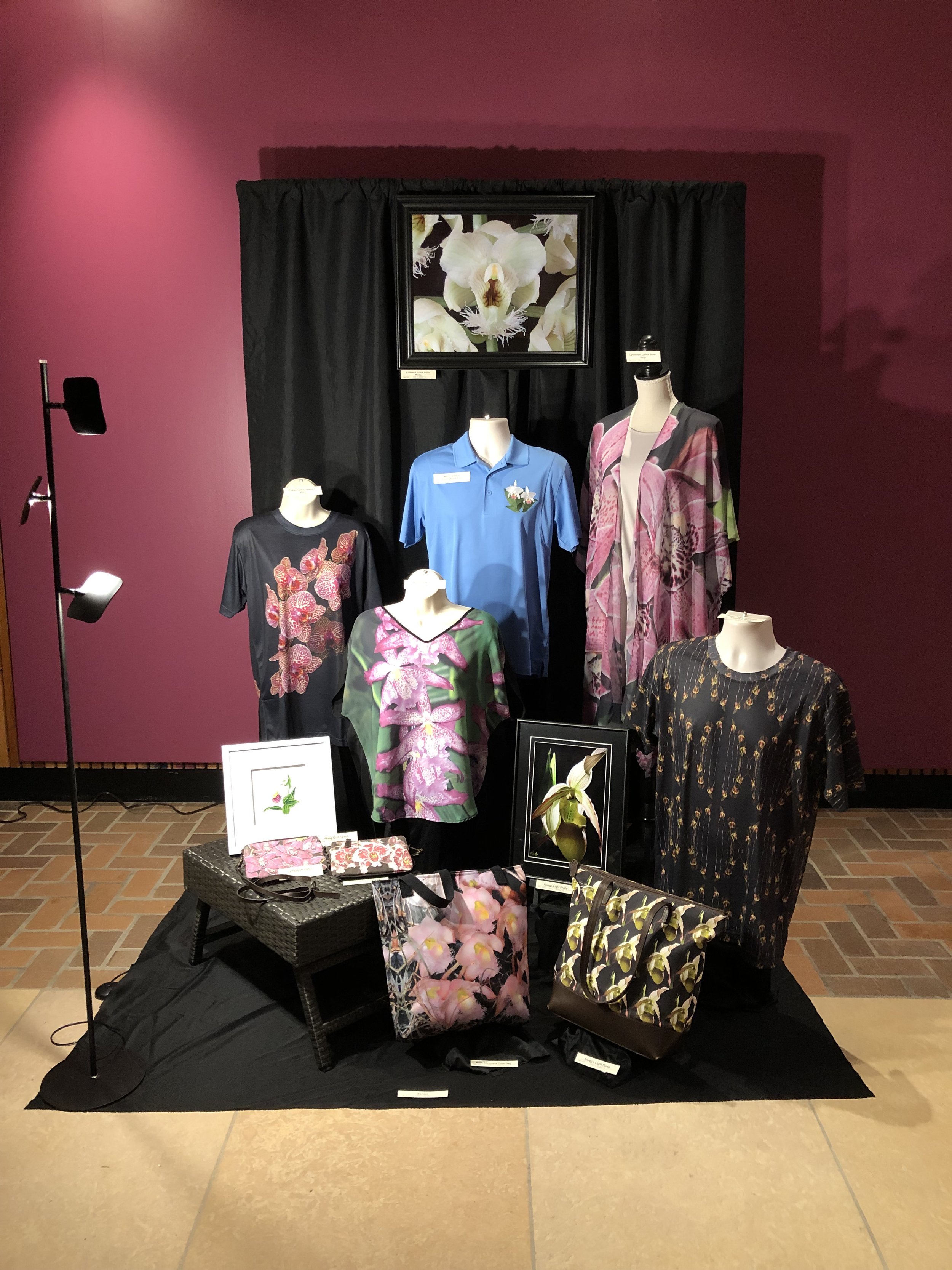 Display of clothing and accessories with floral patterns, including shirts, dresses, and handbags, on a black table and mannequins, against a dark background with a large framed orchid photograph.