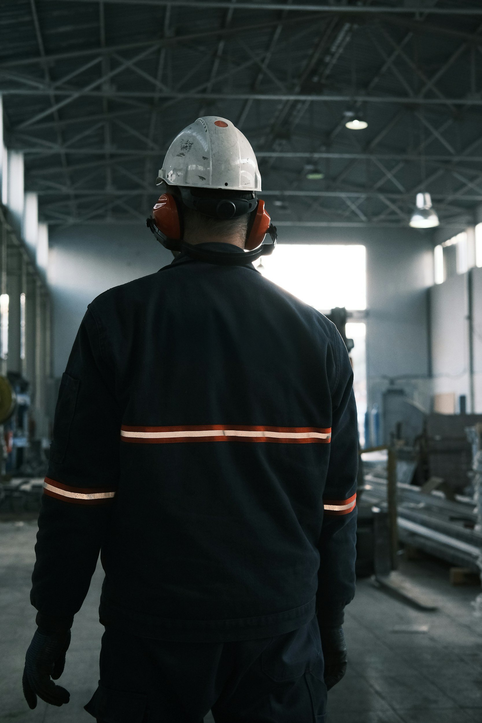 A worker wearing a safety helmet, ear protection, and dark clothing inside an industrial warehouse or factory.