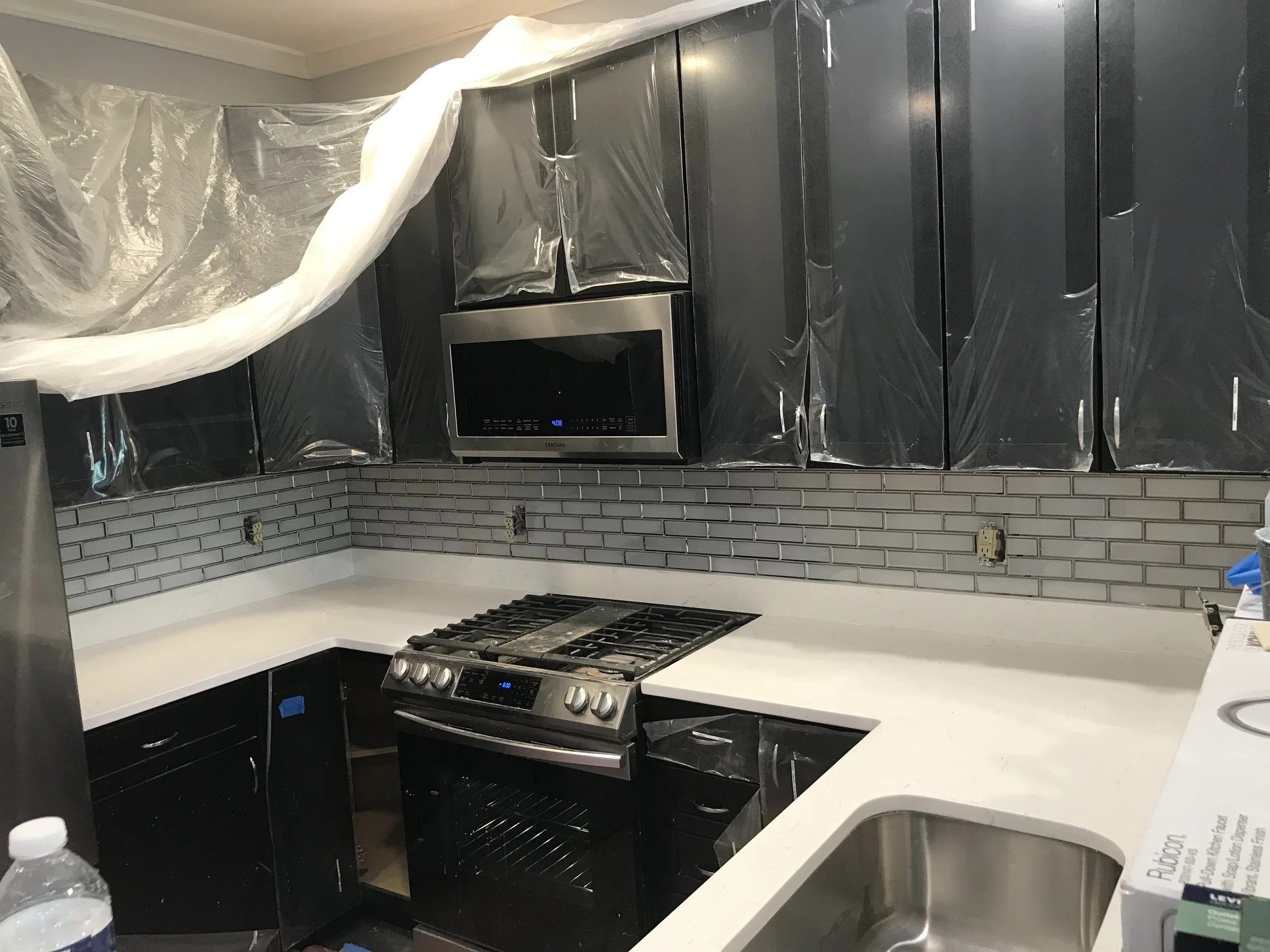 Kitchen with black cabinets covered in plastic, a microwave, and a stove, with a gray subway tile backsplash.