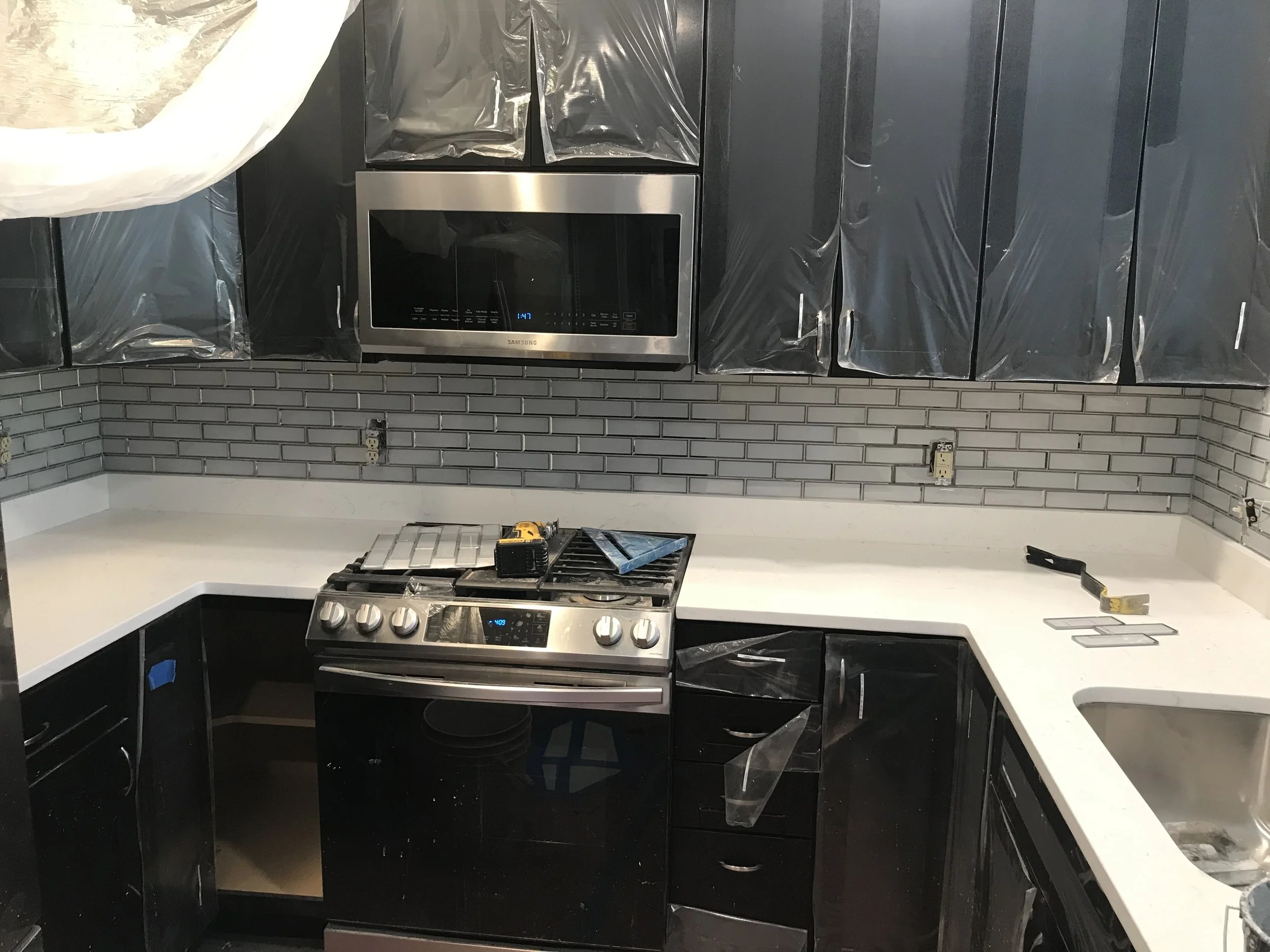 Kitchen with black cabinets, a stainless steel microwave, stovetop, and a gray tile backsplash. Plastic covers are seen on some cabinets and appliances, indicating ongoing installation or renovation.