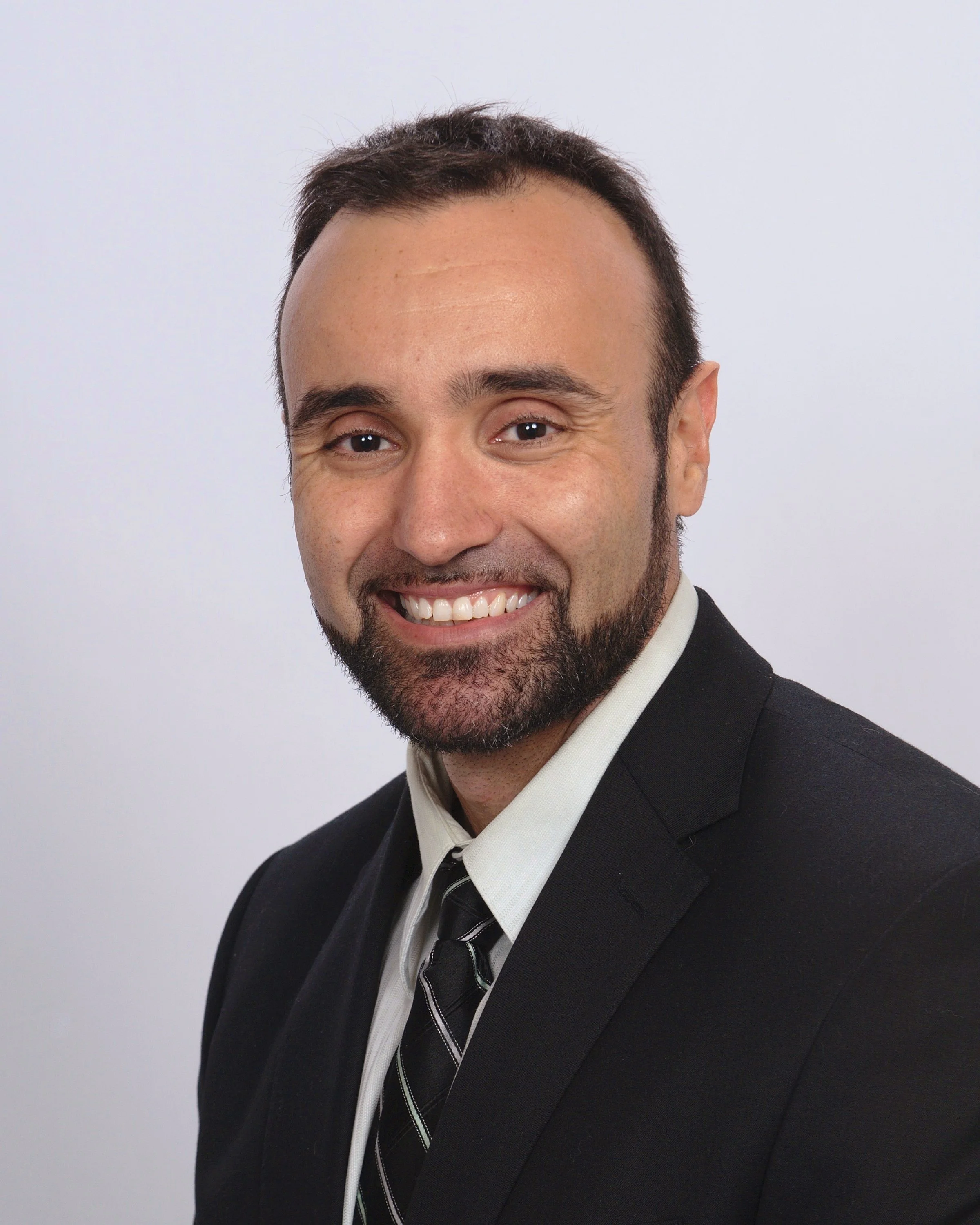 Close-up of a smiling man with dark hair and a beard, wearing a black suit, white shirt, black tie, and a striped tie, against a plain white background.