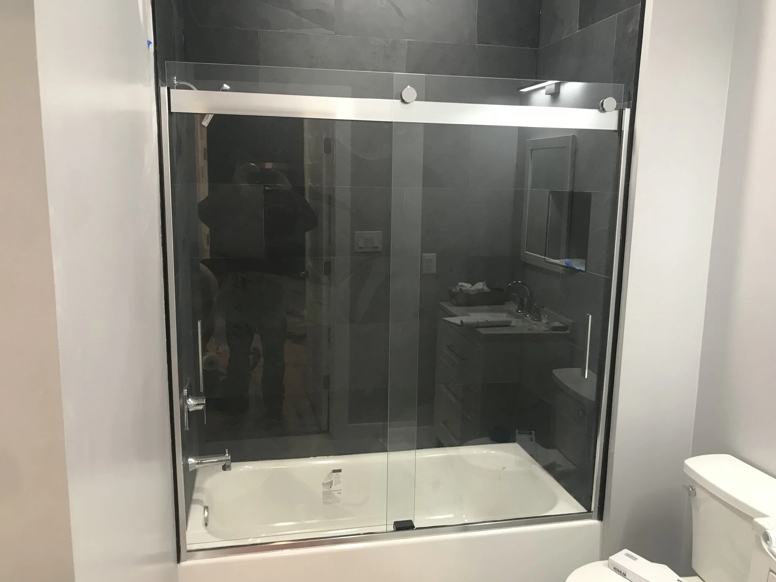 Bathroom with a walk-in shower with black tiles, glass door, and white fixtures, and a toilet nearby.