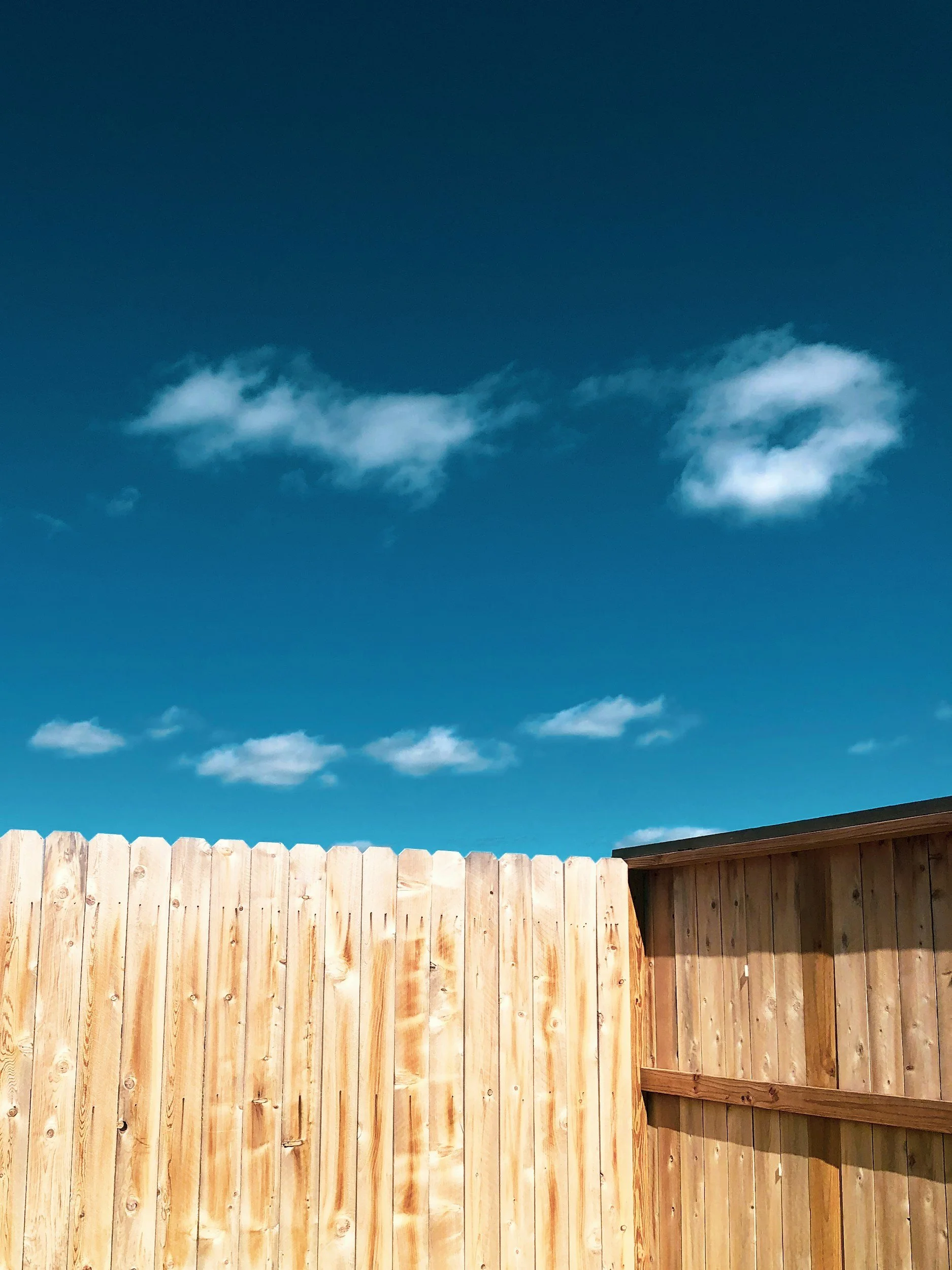 View of a light wooden fence and part of a shed or enclosed structure against a blue sky with scattered white clouds.