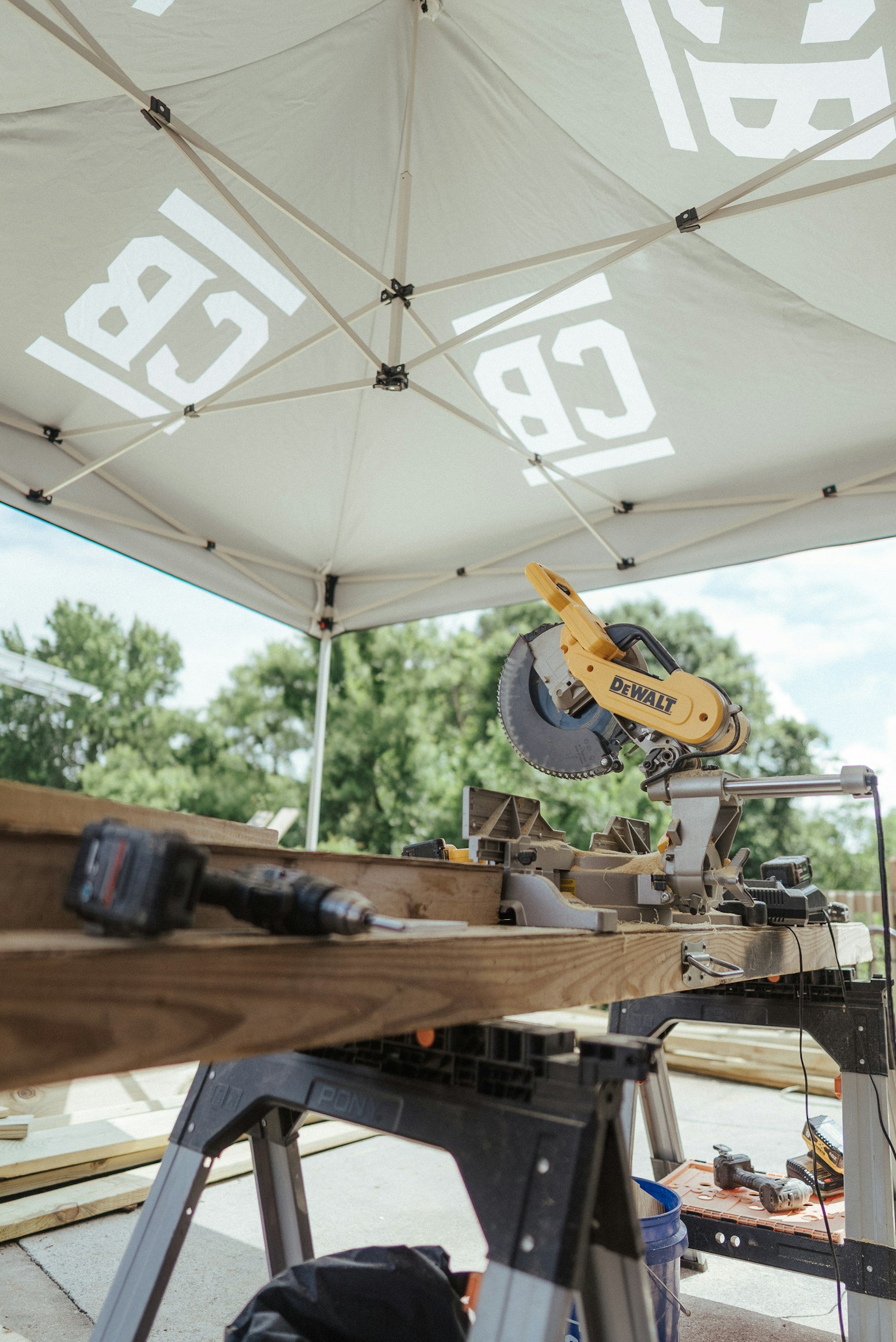 A woodworking area outdoors with a DeWalt miter saw on a wooden workbench, under a white canopy tent, with tools and power drills nearby.