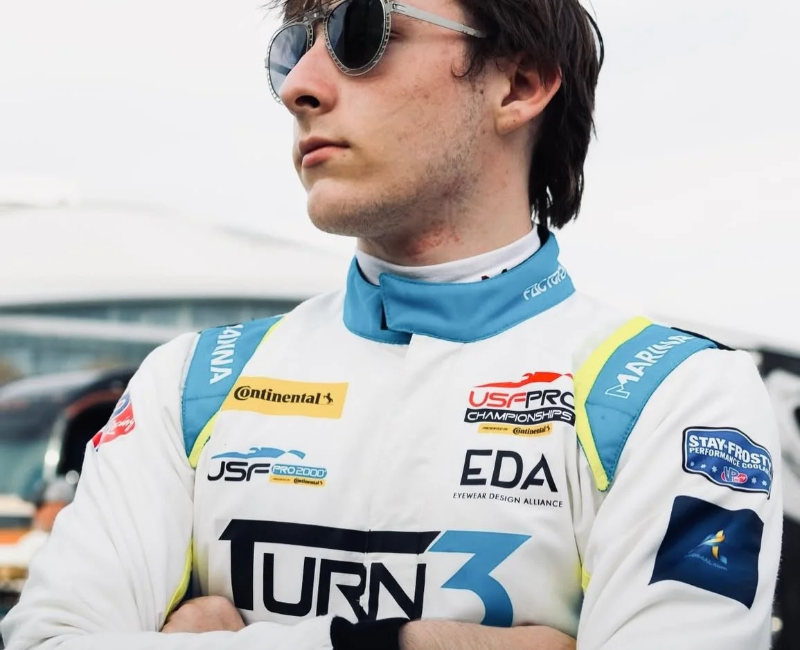 A young race car driver with dark hair wearing sunglasses and a white racing suit with various sponsor logos, standing with arms crossed. Looking good in Dallas Texas!